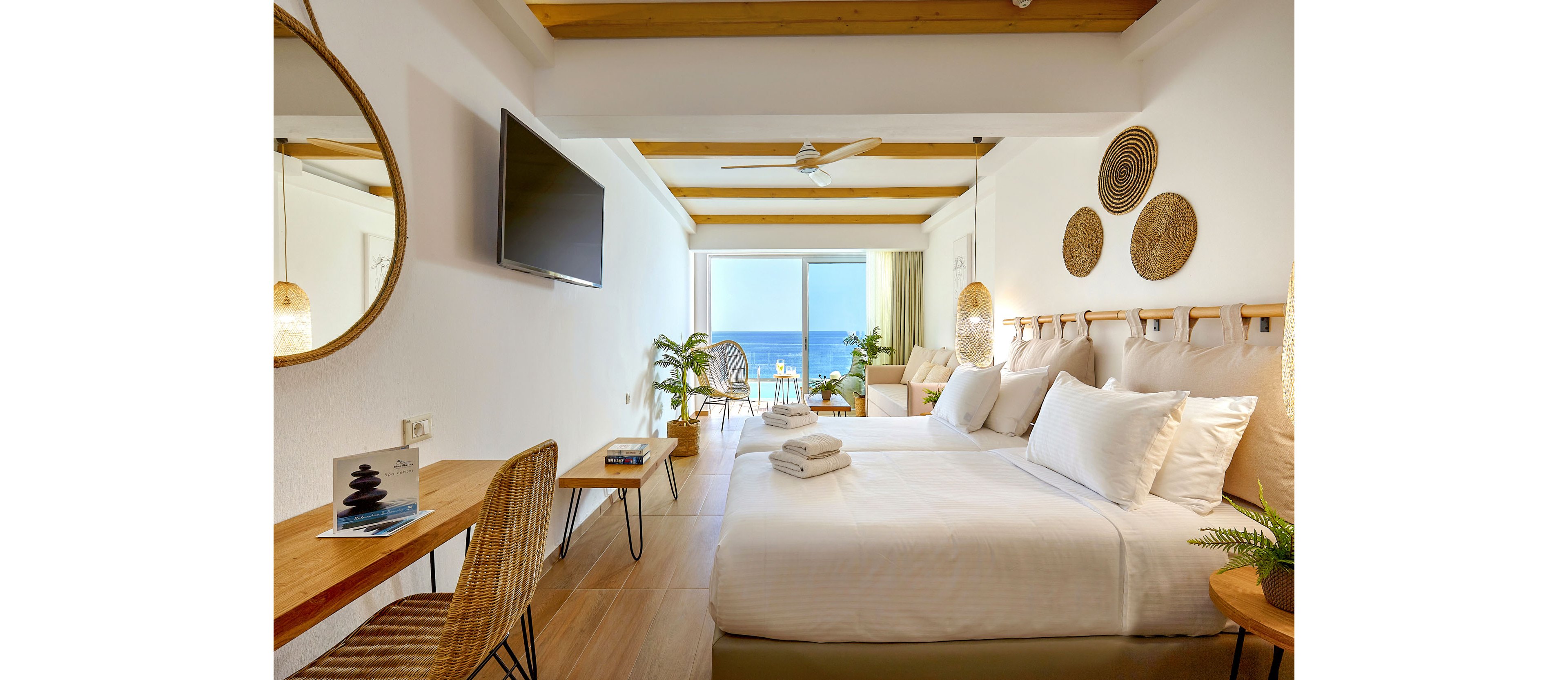Modern hotel room with a large bed and neatly folded white towels in the foreground. The interior features light wood flooring, wicker furniture, and decorative wall art, creating a warm, natural atmosphere. A flat-screen TV is mounted on the wall opposite the bed, and a round mirror and desk area sit to the side. Floor-to-ceiling glass doors open to a balcony with chairs overlooking a clear blue ocean, filling the space with natural light.