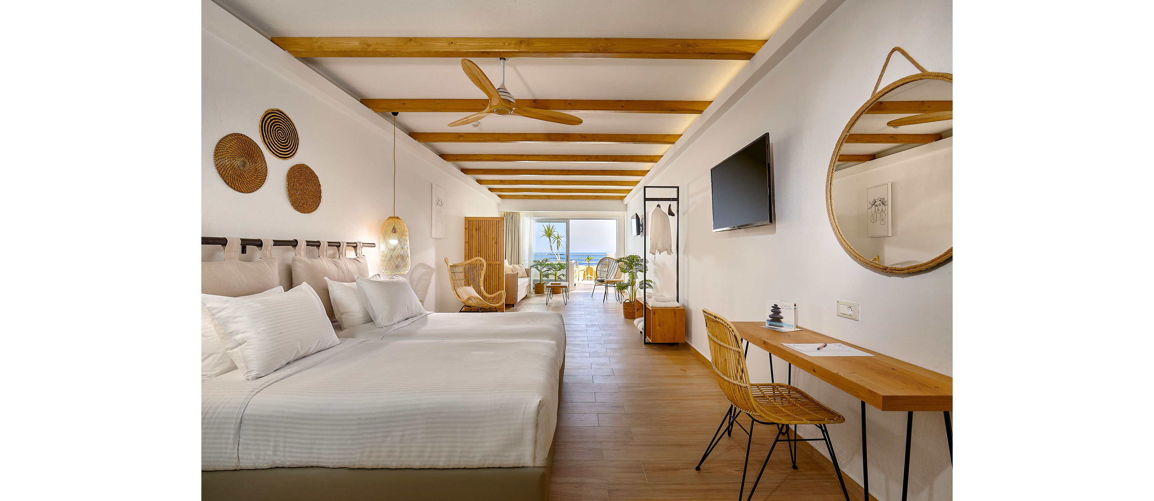 Spacious modern hotel room features a large bed with white linens and multiple pillows. The interior showcases wooden ceiling beams, wicker accents, and a minimalist desk area with a round mirror and chair. A flat-screen TV is mounted on the wall opposite the bed, and decorative baskets and pendant lights add warmth. Floor-to-ceiling glass doors open to a balcony with outdoor seating and a scenic view of palm trees and the sea.