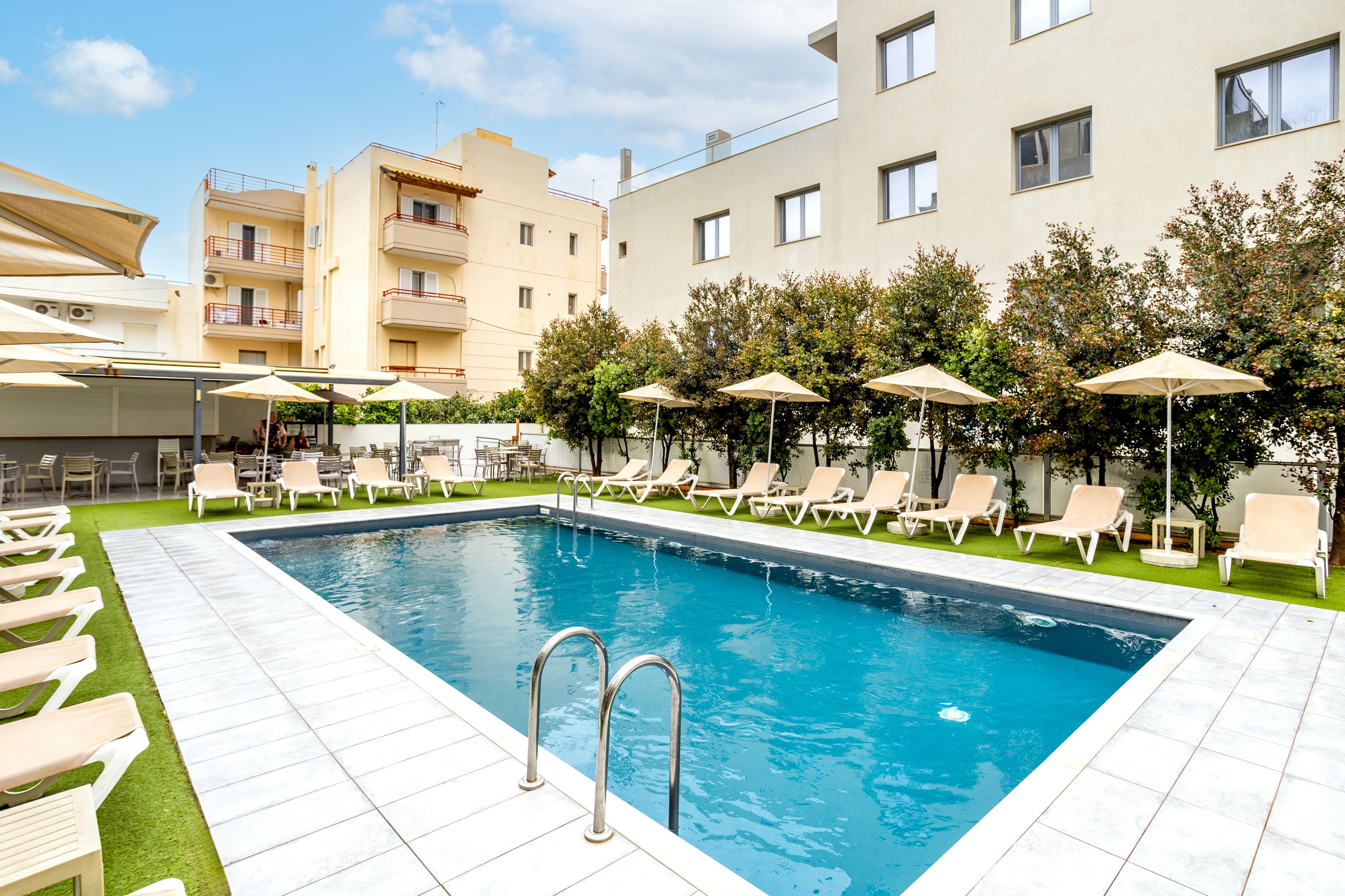 An outdoor rectangular swimming pool is set in the courtyard of a modern hotel or apartment building. Beige lounge chairs and cream-colored umbrellas line the poolside on artificial grass, creating a relaxed resort atmosphere. Low shrubs provide greenery and privacy between the pool area and the surrounding buildings. The sky is bright with scattered clouds, suggesting a pleasant, sunny day.