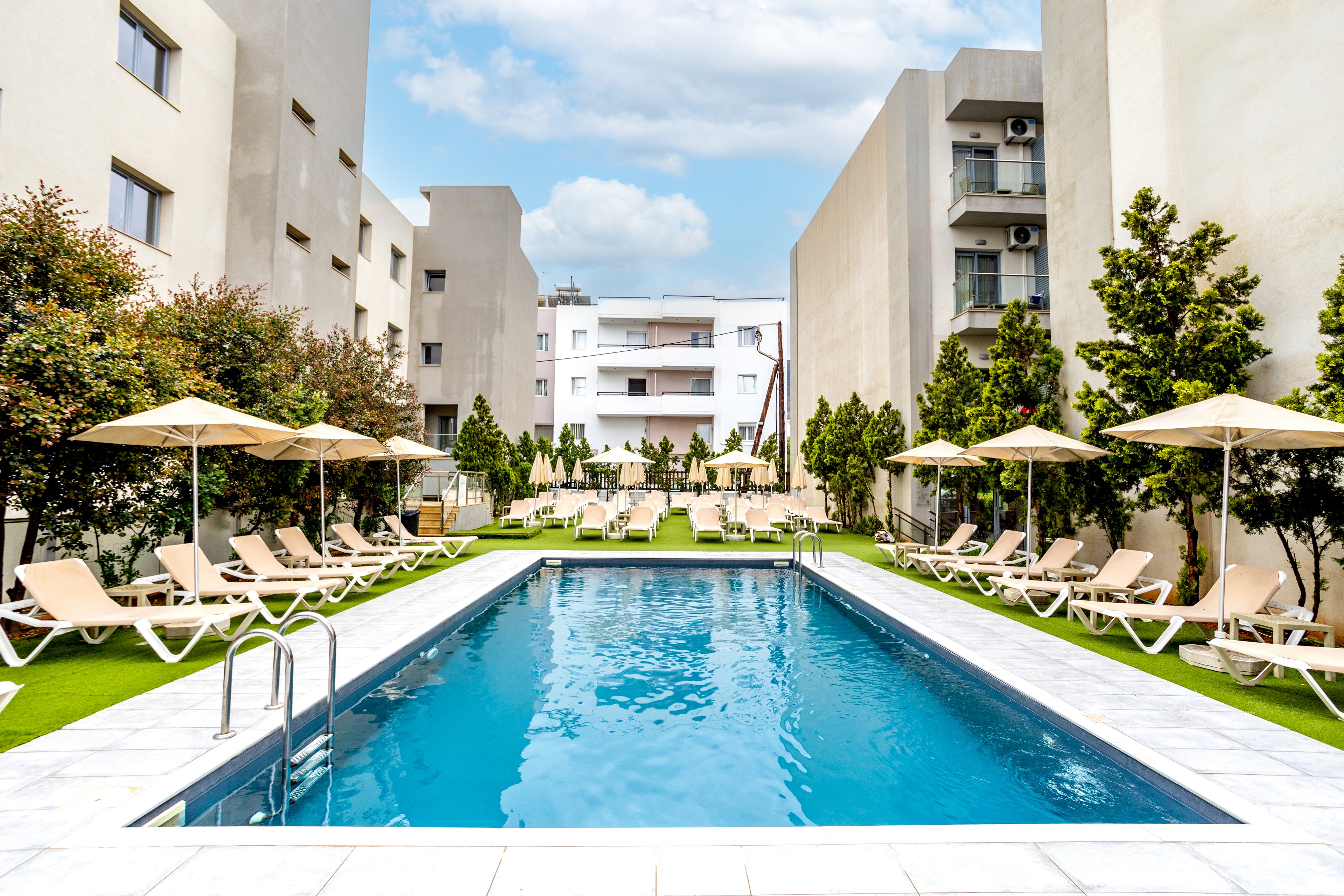 A long rectangular swimming pool is centered in a modern apartment courtyard. Beige lounge chairs and umbrellas line both sides of the pool on bright green artificial grass. Tall residential buildings frame the scene, creating an enclosed, private atmosphere. The sky is partly cloudy, and the overall setting feels clean, bright, and inviting for relaxation.