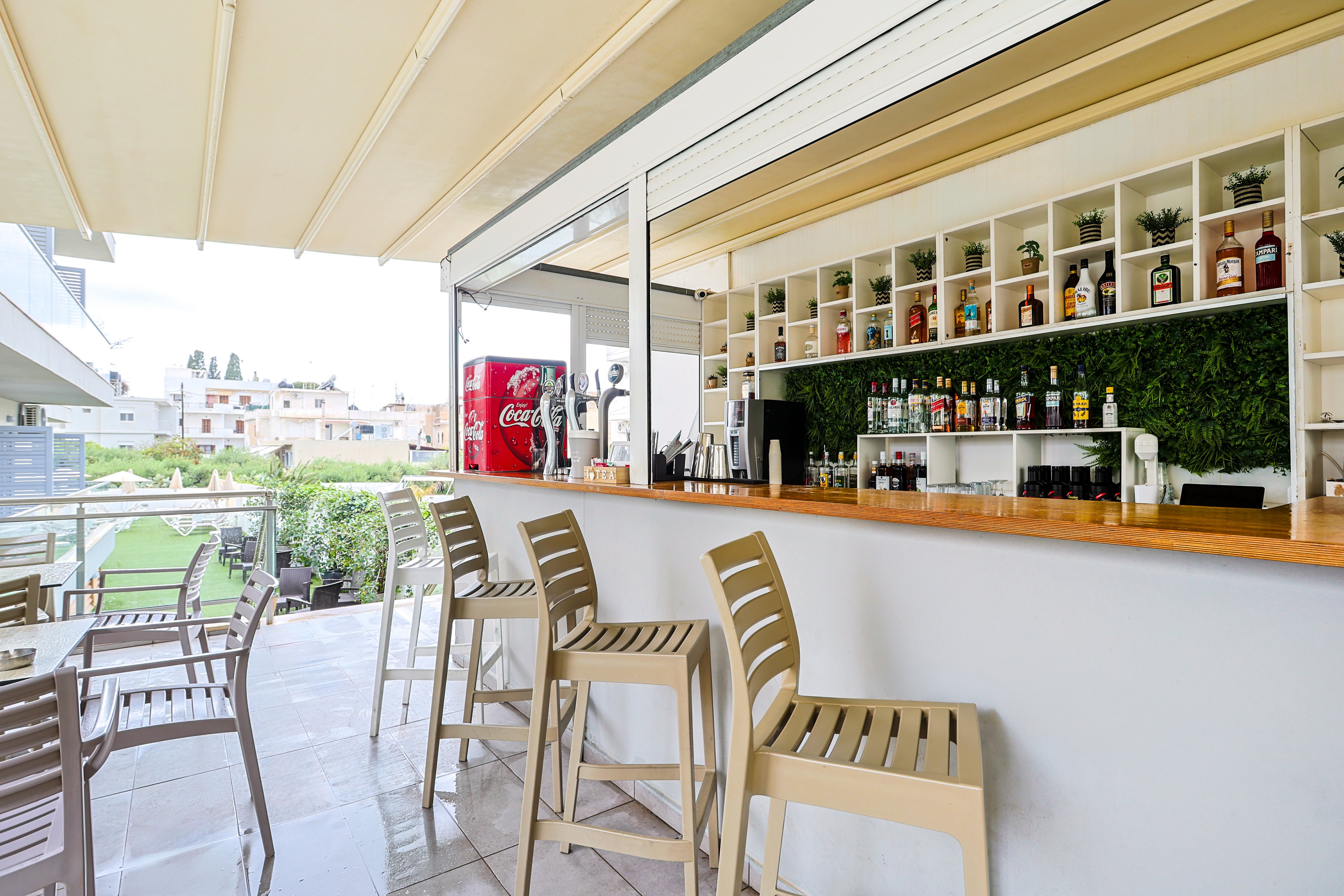 A contemporary outdoor bar features a long wooden counter with several beige bar stools neatly arranged along it. Behind the counter, shelves display a variety of liquor bottles and small potted plants against a green wall backdrop. Additional tables and chairs are set up on the terrace, overlooking a landscaped area and nearby buildings. The space is covered by a light-colored canopy, creating a bright yet shaded atmosphere suitable for daytime or evening drinks.