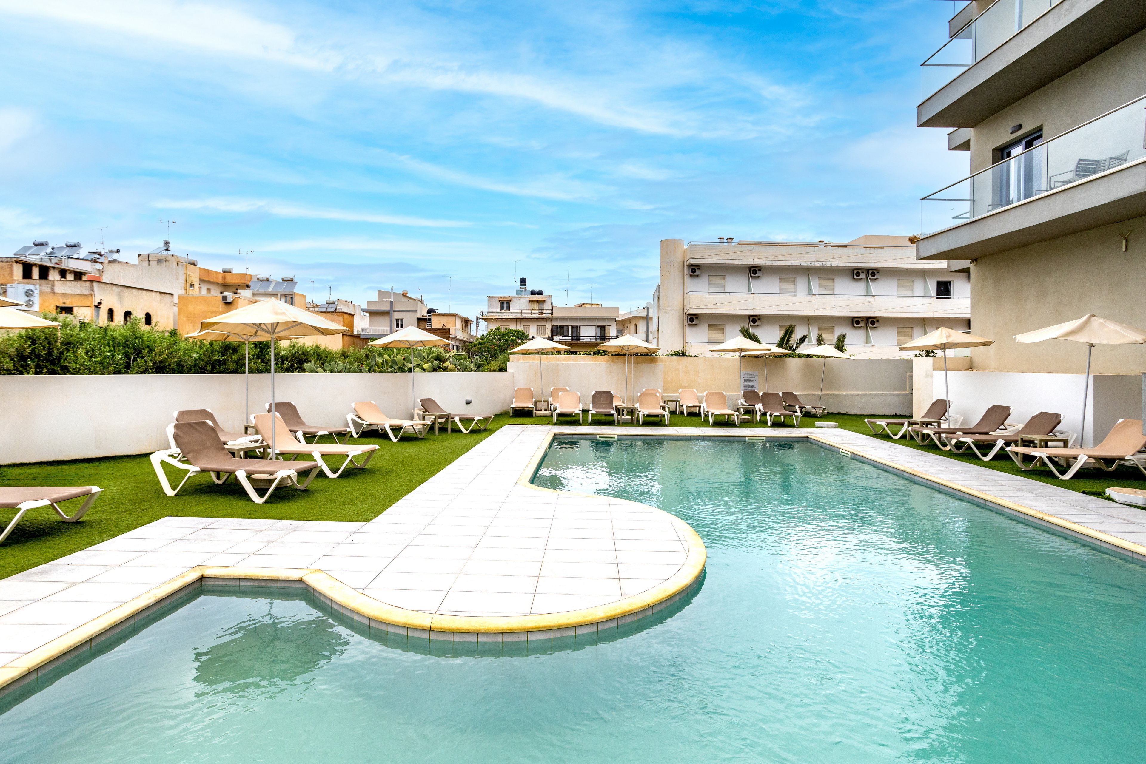 A modern outdoor swimming pool curves around a tiled deck beside a contemporary hotel building. Numerous beige sun loungers and umbrellas are arranged on artificial grass along the poolside, suggesting a relaxing leisure area. Low white walls separate the pool terrace from neighboring residential buildings in the background. The bright blue sky and soft clouds create a calm, inviting atmosphere for guests.
