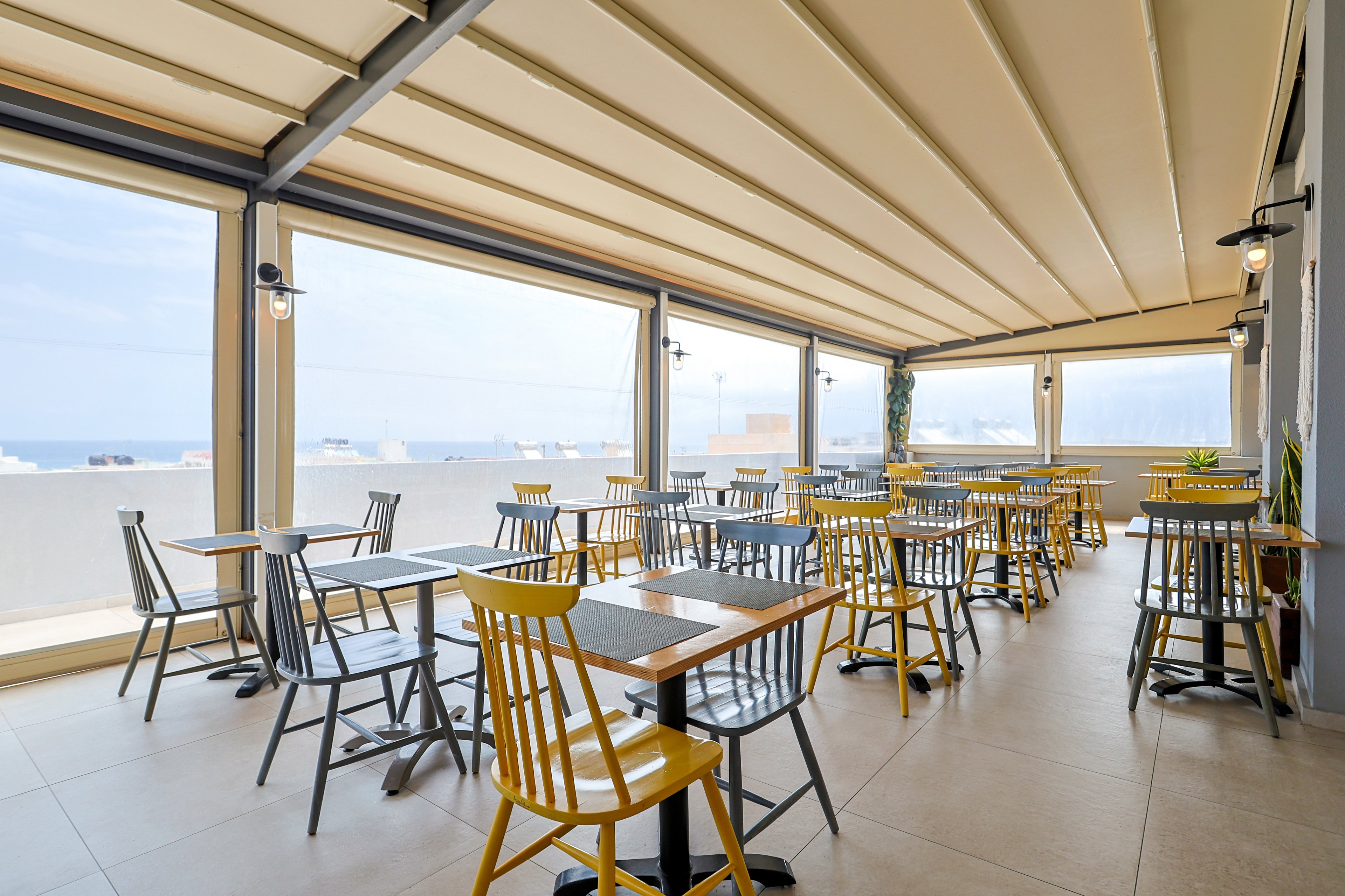 An open-air terrace cafe features neatly arranged wooden tables with yellow and gray chairs. The space is covered by a light-colored retractable roof that filters soft daylight. Large open windows reveal a distant view of the sea and sky beyond the balcony wall. The overall atmosphere is clean, modern, and inviting, ready to welcome guests.