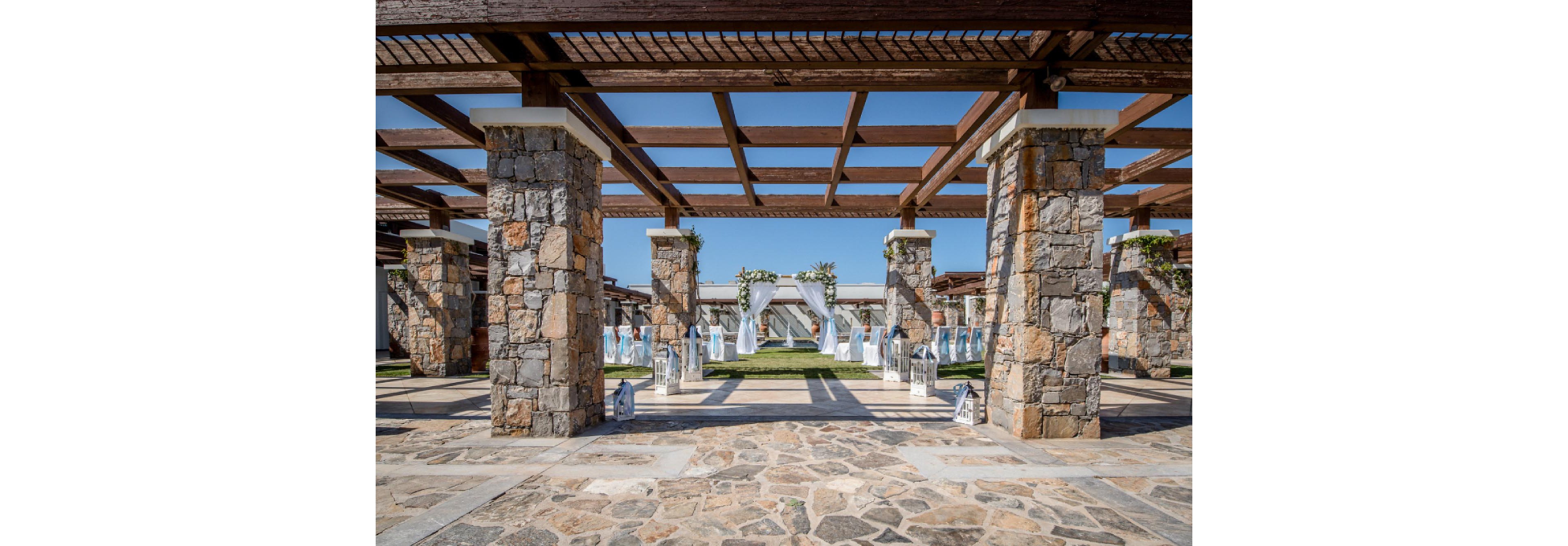 An outdoor wedding ceremony is arranged beneath a large stone pergola with wooden beams. Rows of white chairs line a central aisle leading to a floral archway at the far end. Decorative lanterns are placed along the stone pathway, enhancing the elegant setup. The bright blue sky and distant sea create a scenic, open-air backdrop for the event.