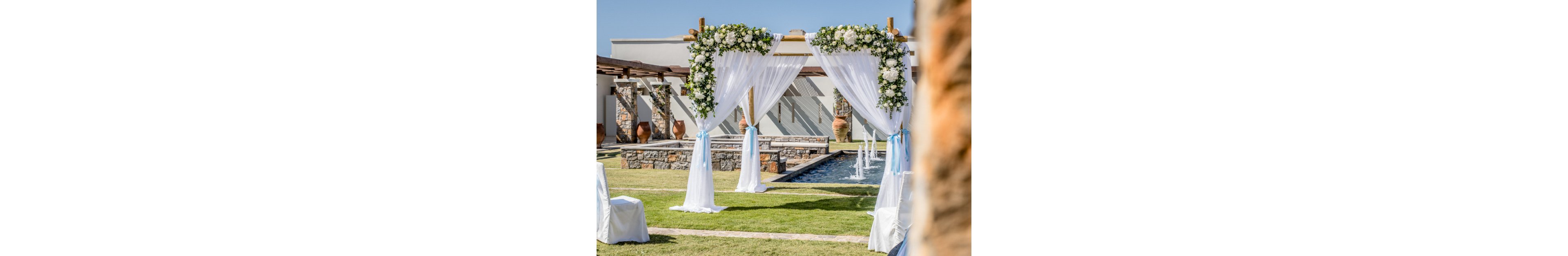 An elegant outdoor wedding ceremony setup features a floral arch draped in white fabric. The arch is decorated with lush white and green flower arrangements and light blue ribbons. In the background, a stone fountain with multiple water jets adds a sophisticated touch to the venue. White covered chairs are arranged on the green lawn, suggesting a formal event under clear blue skies.