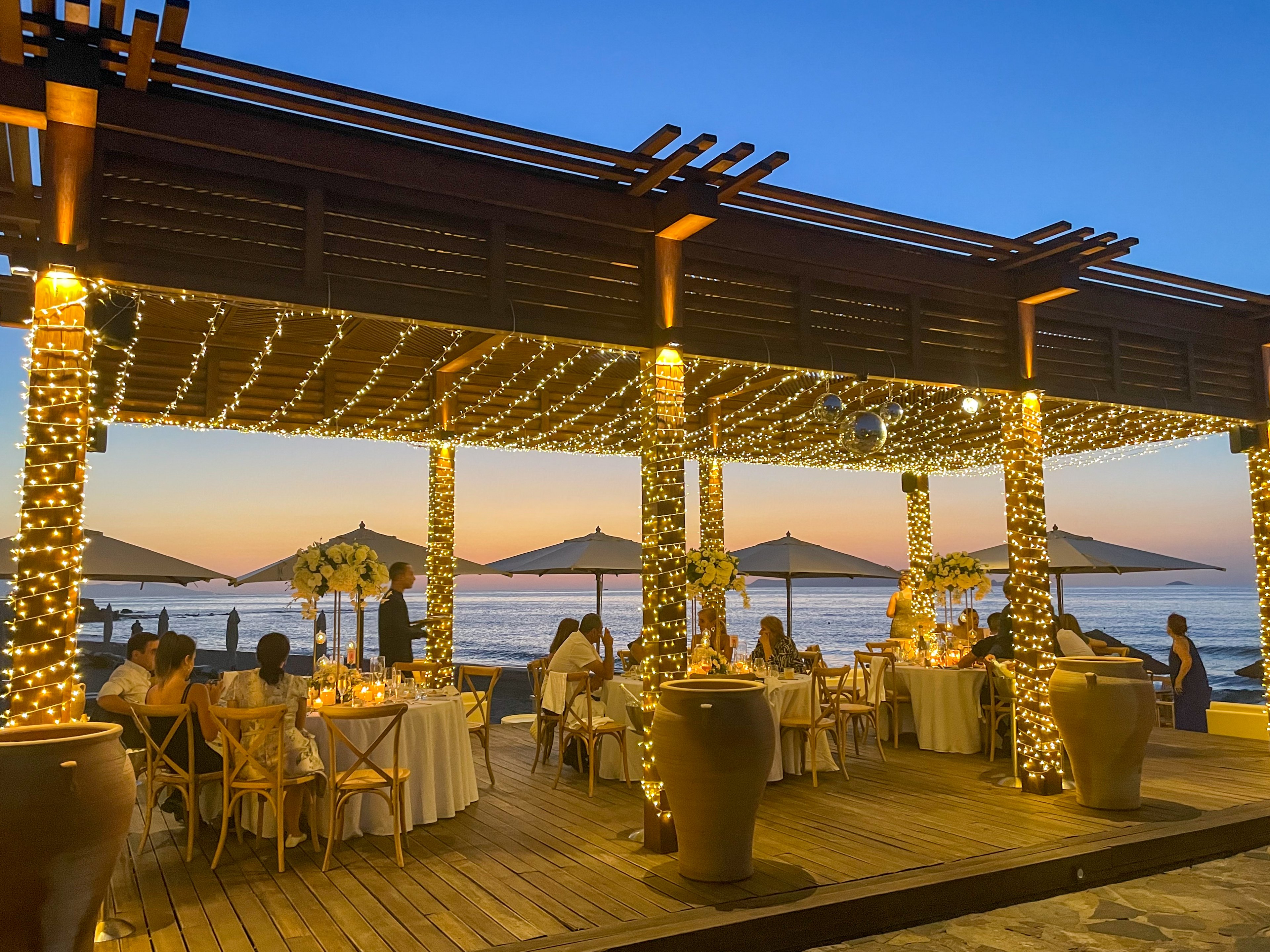 An elegant open-air dining area is set up on a wooden deck by the sea at sunset. Guests sit at round tables with white linens and floral centerpieces, enjoying an intimate meal. The pergola structure is wrapped in warm string lights, creating a cozy, festive glow. Large vases and beach umbrellas frame the scene, while the calm ocean and colorful sky provide a serene backdrop.