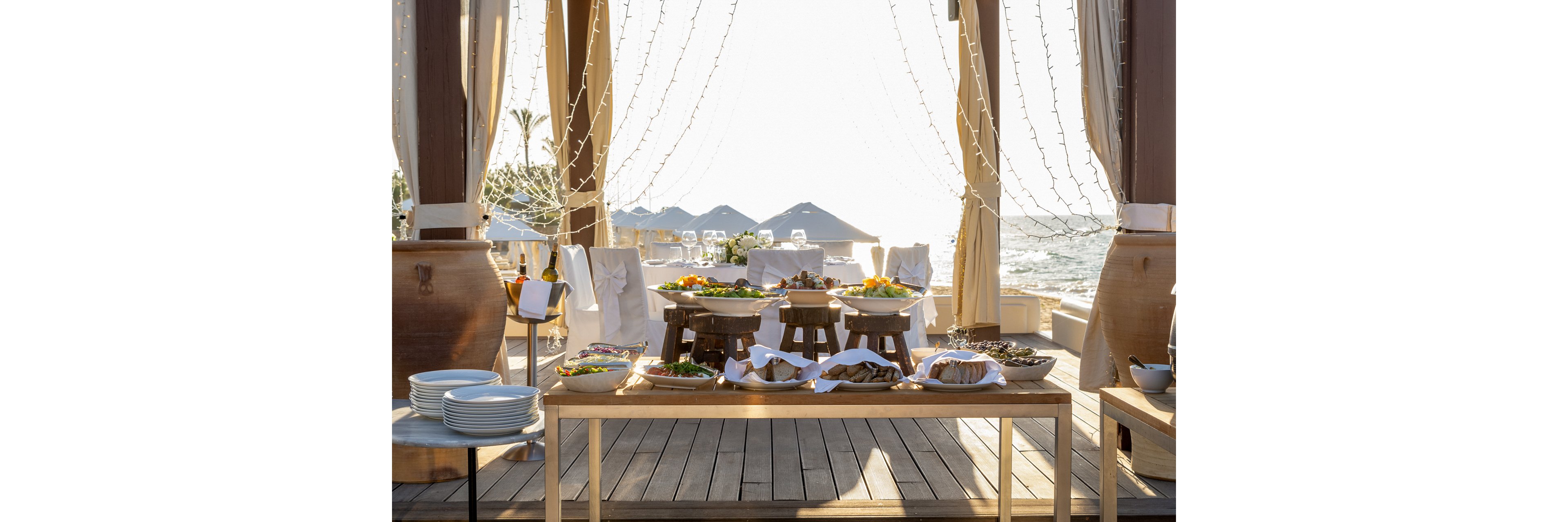 A long wooden table is arranged with an assortment of dishes for a buffet by the sea. Covered serving bowls and platters sit on raised stands, surrounded by neatly stacked plates and cutlery. Sheer drapes, string lights, and large decorative vases frame the open-air dining area. In the background, sunlit water, umbrellas, and lounge chairs create a relaxed coastal atmosphere, suggesting a festive outdoor event.