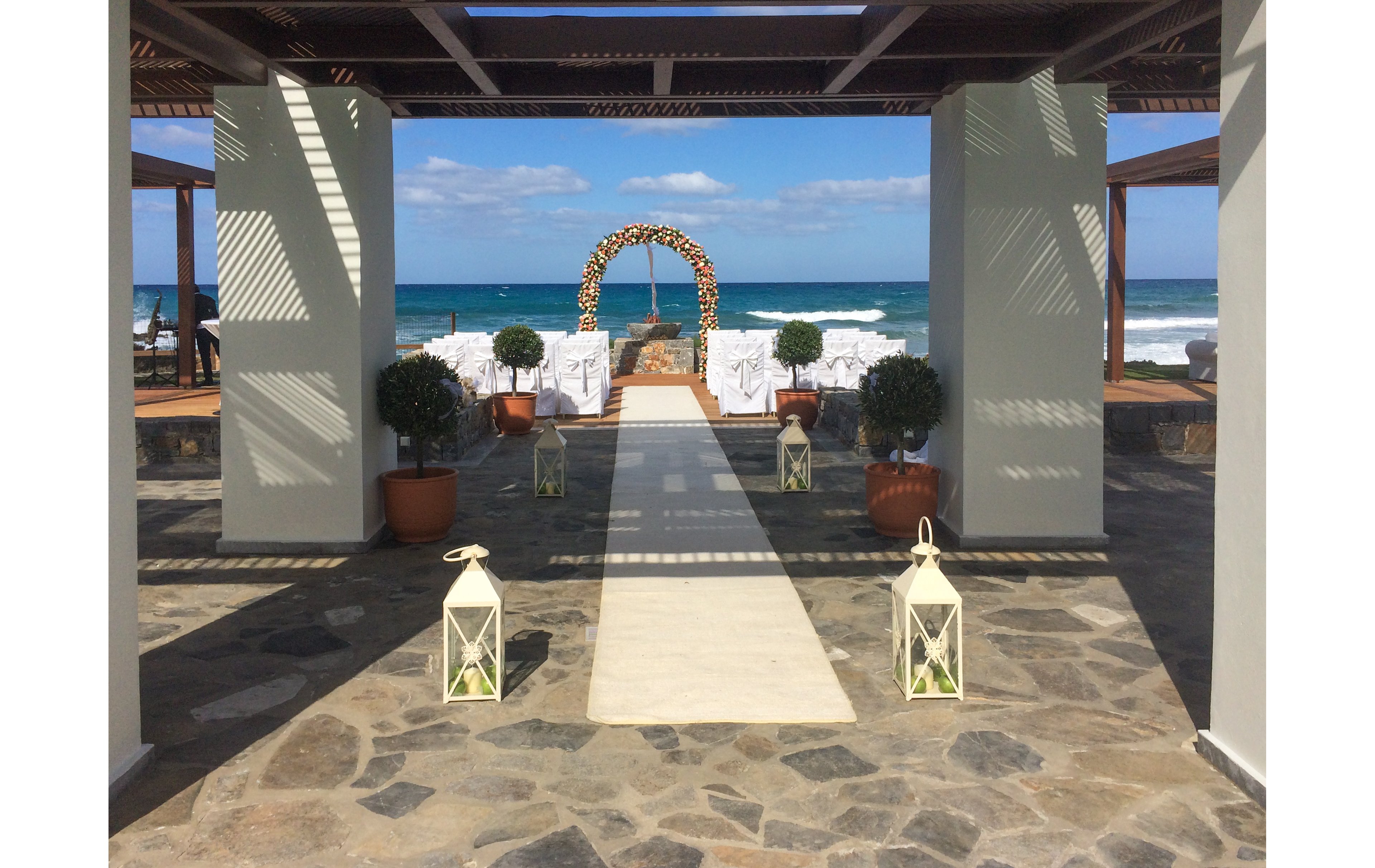 An elegant wedding aisle leads toward a floral archway set against a bright blue ocean backdrop. White lanterns line the stone pathway, creating a romantic entrance framed by potted topiary trees. Rows of white-covered chairs are arranged for guests, facing the ceremony area by the water. The open-air structure and clear sky give the scene a sunny, coastal atmosphere.