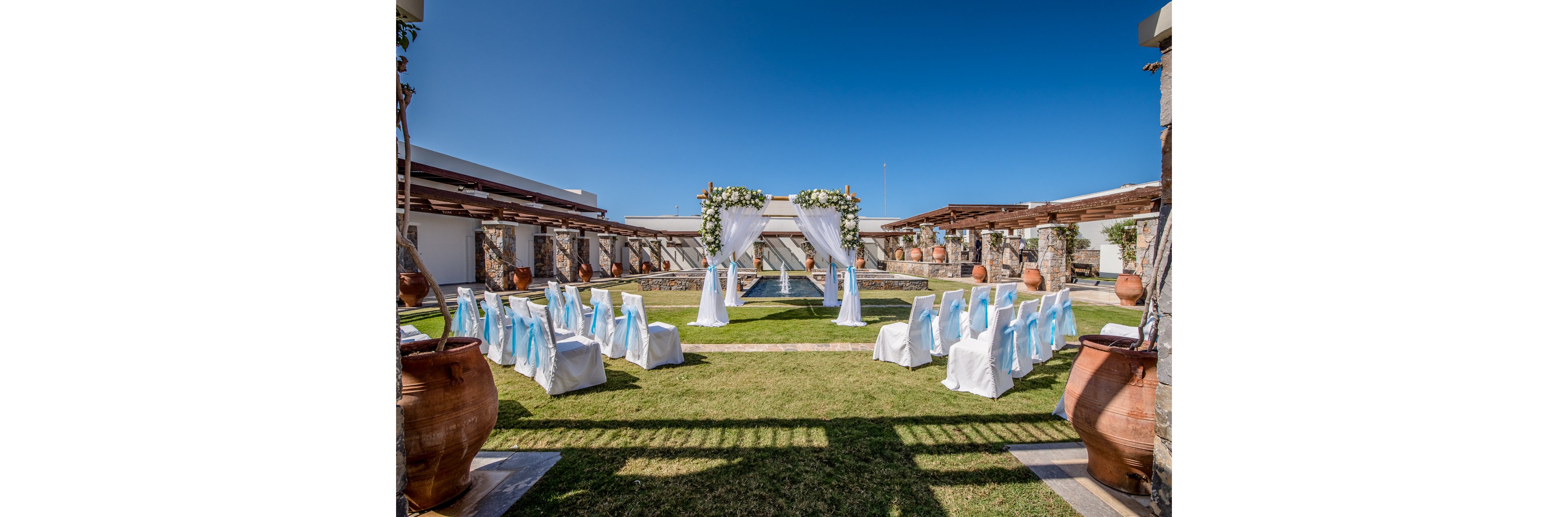 An outdoor wedding ceremony area is arranged around a floral archway and draped white fabric. Rows of white chairs with light blue sashes form an aisle leading to the altar structure over a small water feature. The venue is surrounded by stone columns, wooden pergolas, and large terracotta pots. Bright sunlight and a clear blue sky create a festive, romantic atmosphere.