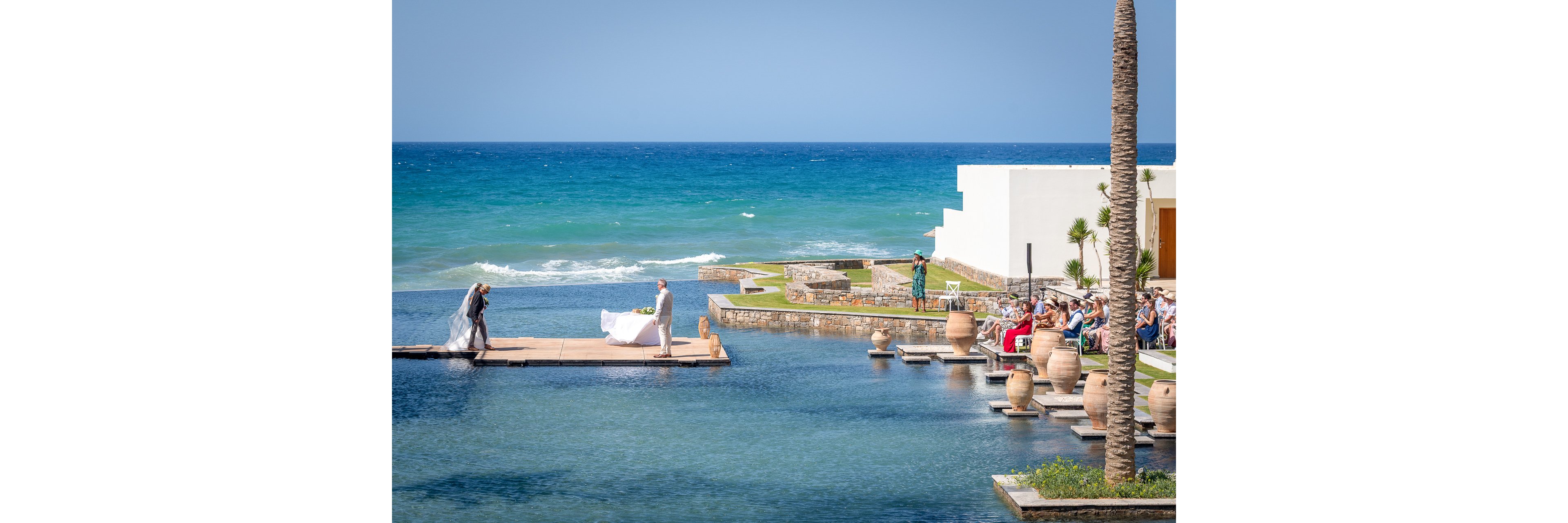 A bride and groom stand on a platform over an infinity pool during an outdoor wedding ceremony by the sea. Guests sit on the right side, watching the couple with the ocean stretching out in the background. The modern resort architecture features white buildings, stone walls, and decorative clay pots along the water. Bright daylight and clear blue skies create a serene, celebratory atmosphere.