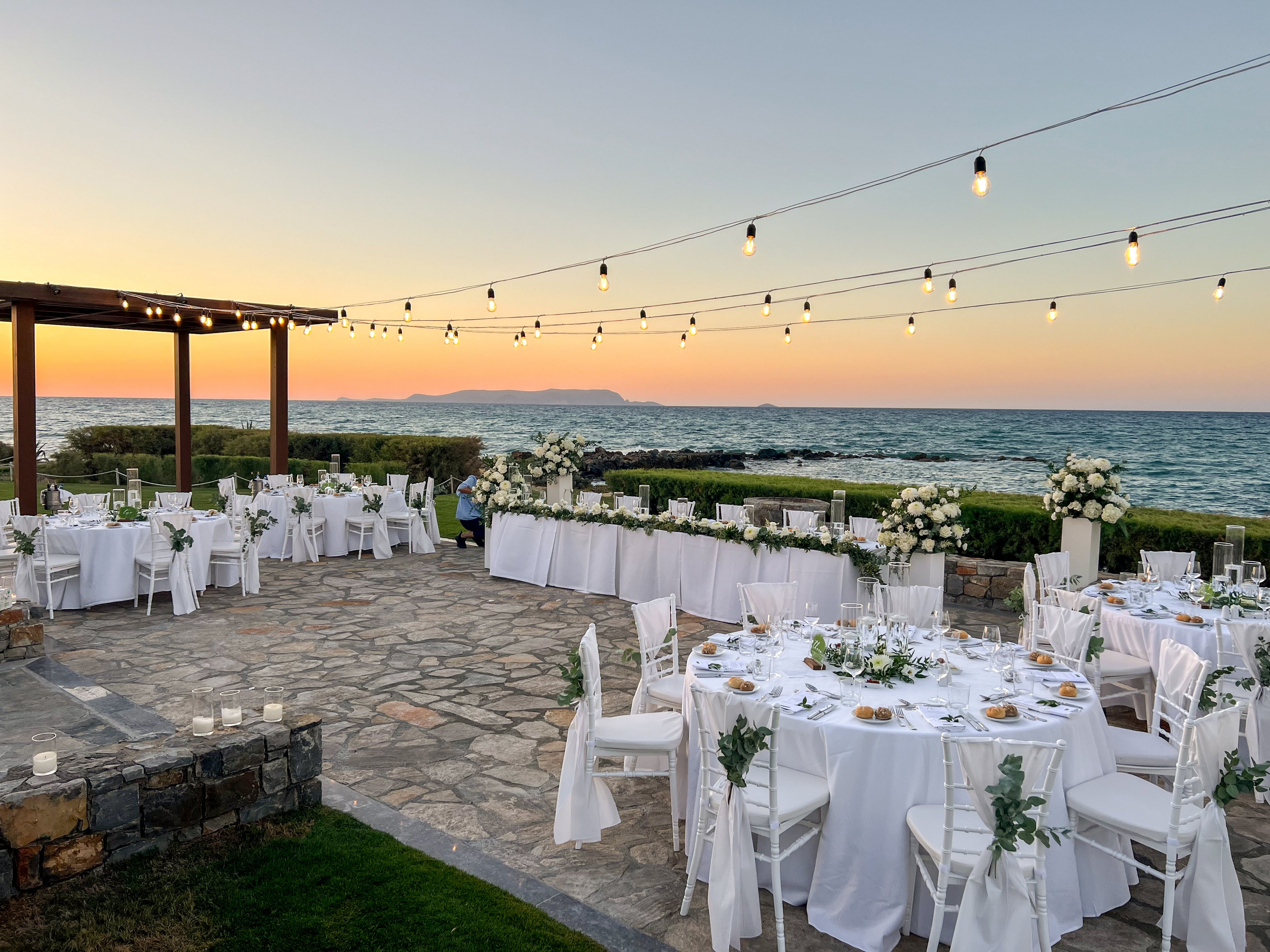 An outdoor wedding reception is set up beside the sea at sunset, with round tables dressed in white linens and floral centerpieces. String lights hang overhead, creating a warm and inviting atmosphere as the sky glows in pastel colors. A long head table is decorated with abundant white flowers and greenery near the water's edge. The stone patio, scattered candles, and ocean horizon complete the romantic coastal setting.