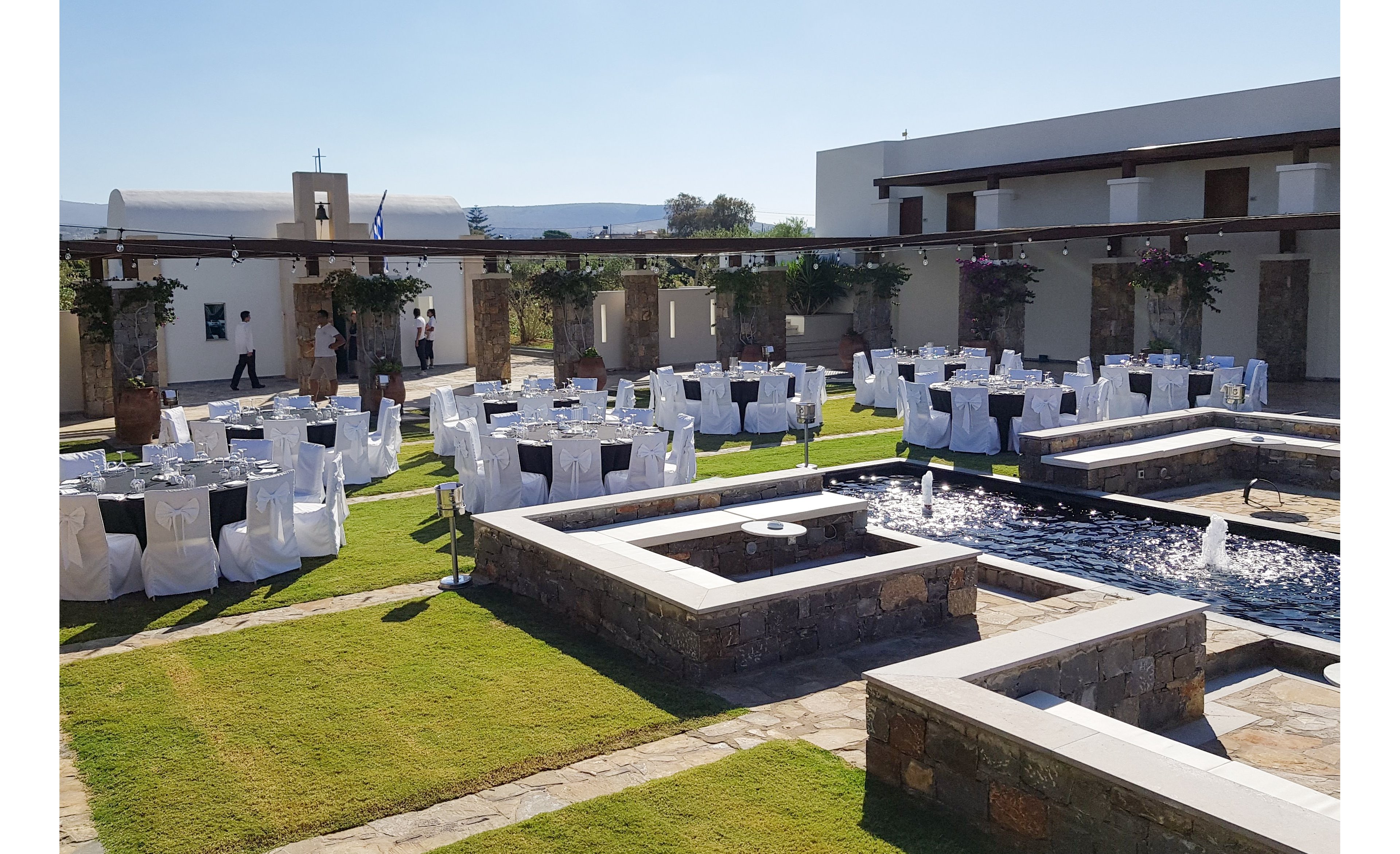 An outdoor courtyard is arranged with round tables covered in white linens and chairs draped in white covers. The setting appears to be part of a modern Mediterranean-style venue with stone walls and white buildings. A central water feature with fountains runs through the middle of the courtyard, bordered by low stone planters and green grass. The scene is brightly lit by daylight, suggesting preparation for a formal celebration or reception.