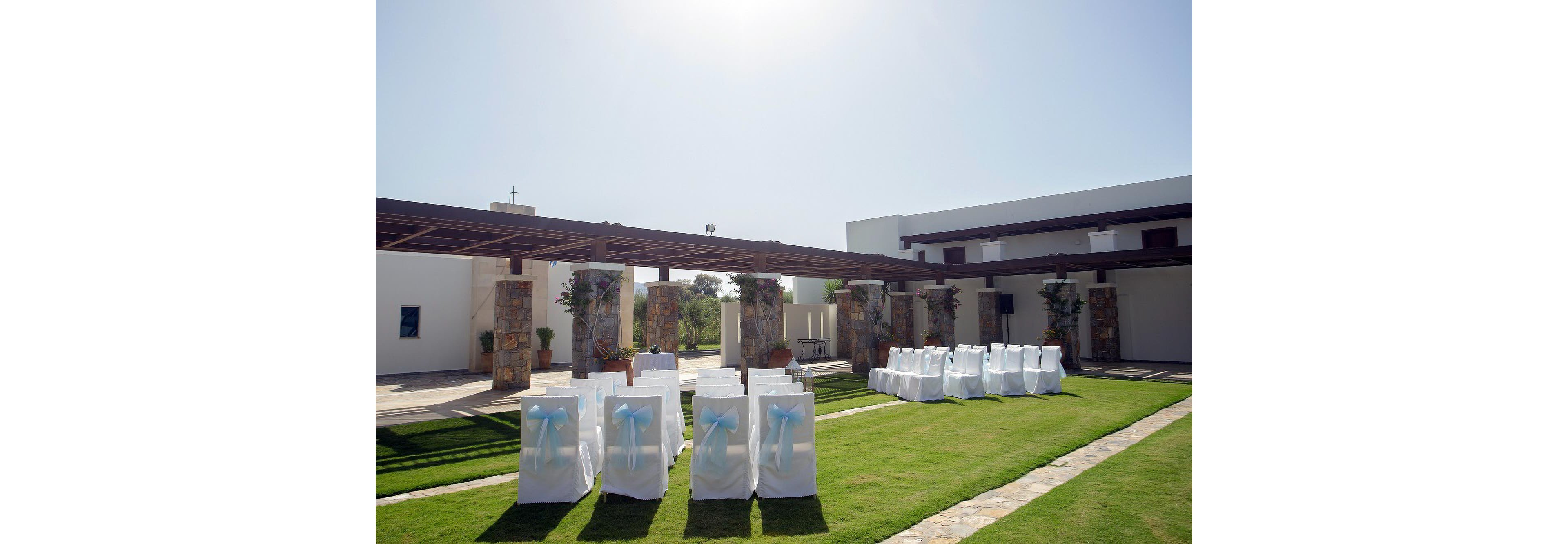 An outdoor wedding ceremony area is arranged on a manicured lawn beside a modern white building. Rows of white chairs with light blue bows are neatly aligned facing a small altar space. A wooden pergola with stone pillars frames the seating area, creating a shaded walkway. The scene is brightly lit under a clear blue sky, suggesting a sunny daytime event.