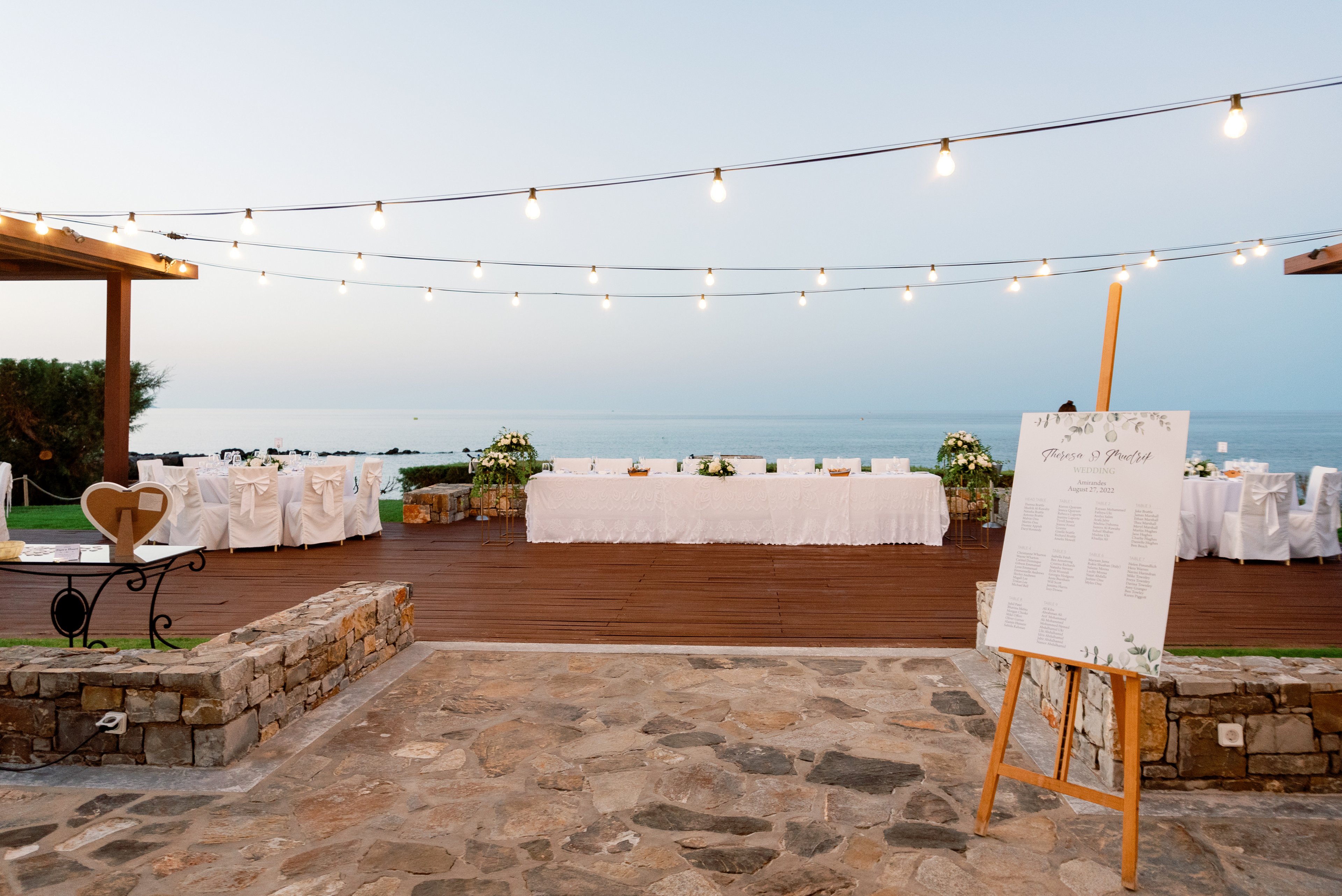 An outdoor wedding reception is set up on a stone and wooden terrace overlooking a calm sea. A long table decorated with white linens and floral arrangements stretches across the deck, framed by string lights hanging overhead. In the foreground, a large seating chart on an easel welcomes guests to the event. Soft evening light and pastel sky tones create a romantic, serene atmosphere.