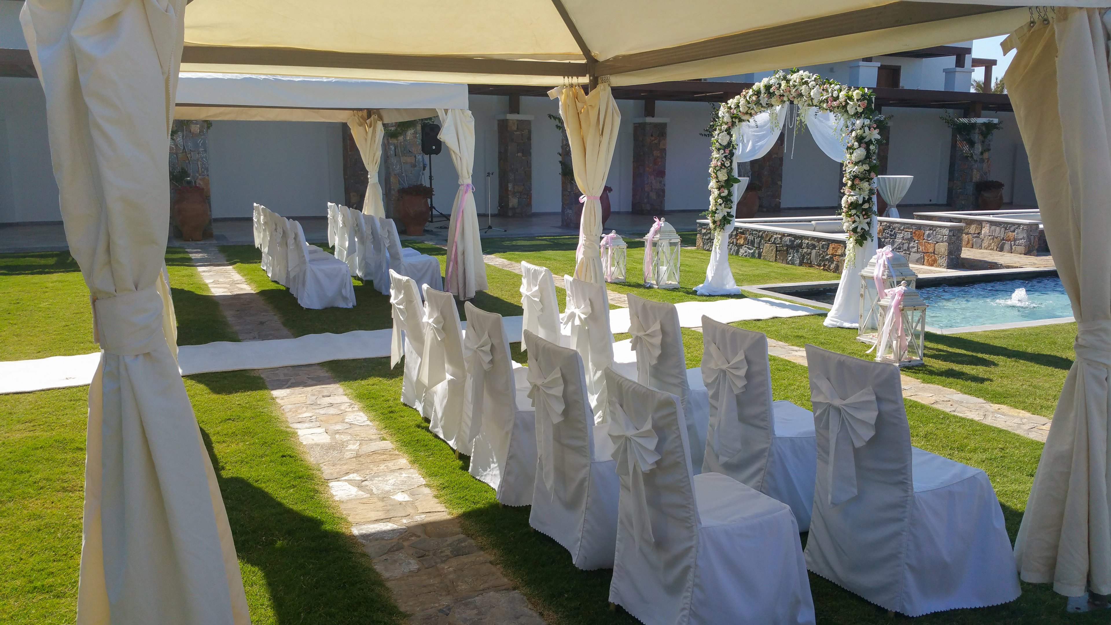 An outdoor wedding ceremony area is arranged with rows of white chair covers tied with bows under a canopy. The chairs face a floral arch positioned near a decorative water feature. Sunlight casts shadows across the green lawn and stone pathway leading to the altar. The scene is elegant and serene, prepared for guests to arrive.