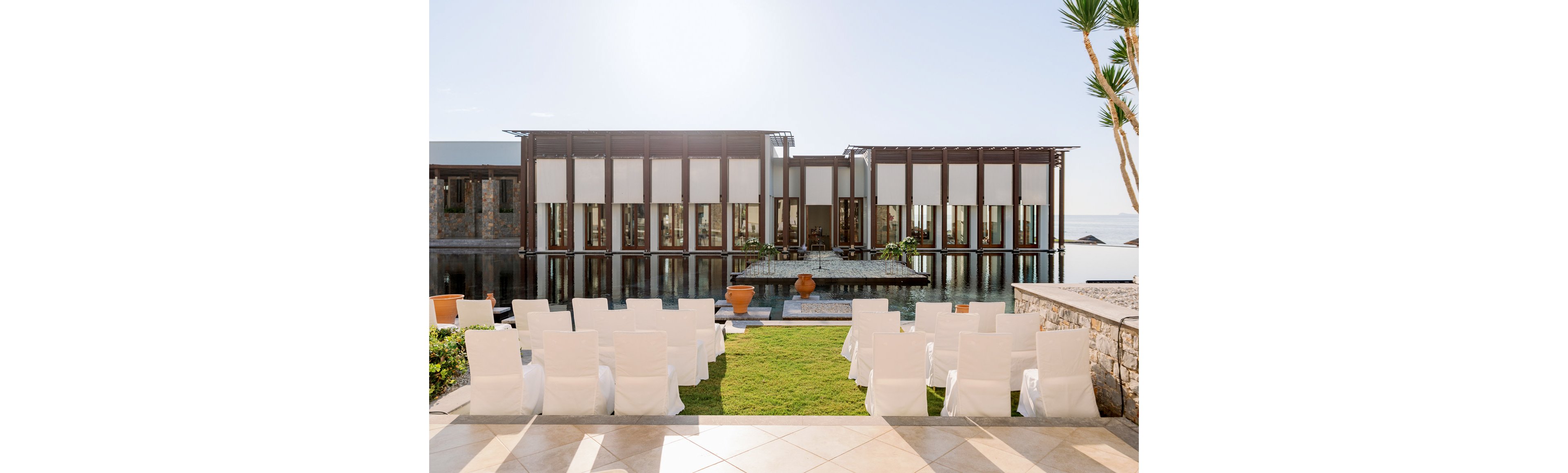 An elegant outdoor ceremony area is arranged with rows of white-covered chairs facing a modern waterfront building. The structure features large glass panels and vertical wooden beams reflected in a calm pool of water. Green lawn, stone walls, and potted plants add natural accents to the contemporary architecture. The bright daylight and clear sky create a serene, romantic atmosphere suitable for weddings or special events.