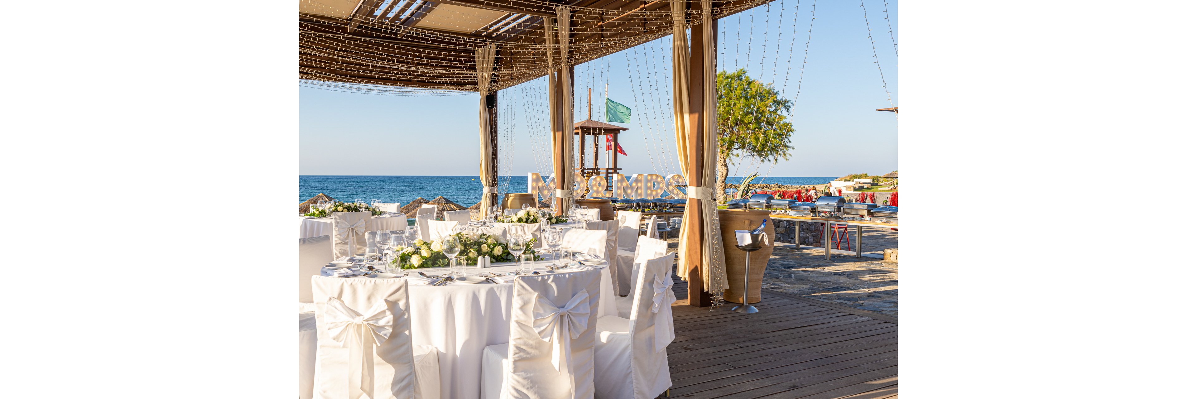 An elegant wedding reception is set up on a wooden deck overlooking the sea. Round tables are covered with white linens, floral centerpieces, and neatly arranged glassware and plates. White chairs with decorative bows surround the tables under a pergola draped with string lights. In the background, a lifeguard tower, flags, and beach seating add to the relaxed coastal atmosphere.