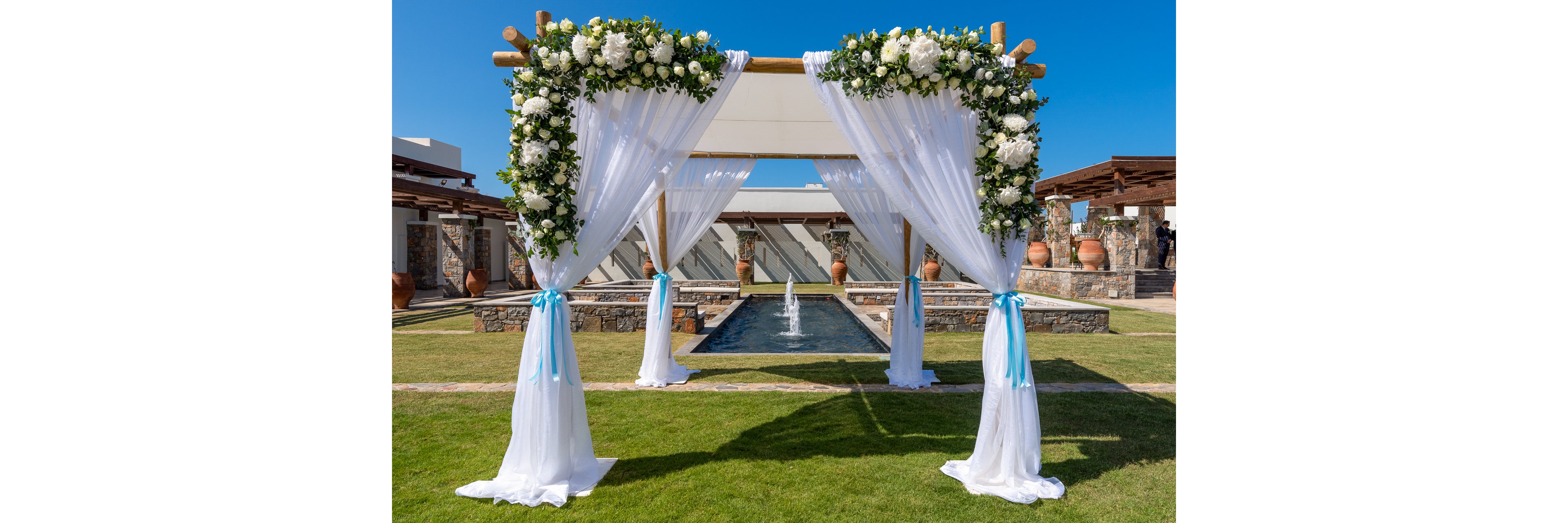 An elegant wooden wedding arch is decorated with flowing white drapes and lush white floral arrangements. The structure stands on a manicured green lawn in front of a long reflecting pool with a central water fountain. Mediterranean-style buildings and large terracotta pots frame the background under a clear blue sky. The scene suggests a romantic outdoor ceremony setup in a sunny, resort-like venue.