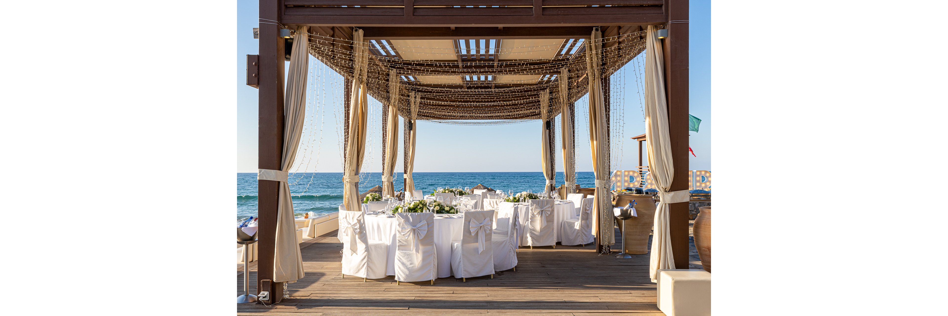 An elegant wedding reception is set beneath a wooden pergola overlooking the sea. Round tables with white linens and chairs tied with bows are arranged on a wooden deck. Sheer curtains and hanging string lights decorate the structure, creating a romantic atmosphere. The calm ocean and clear sky in the background enhance the serene coastal setting.