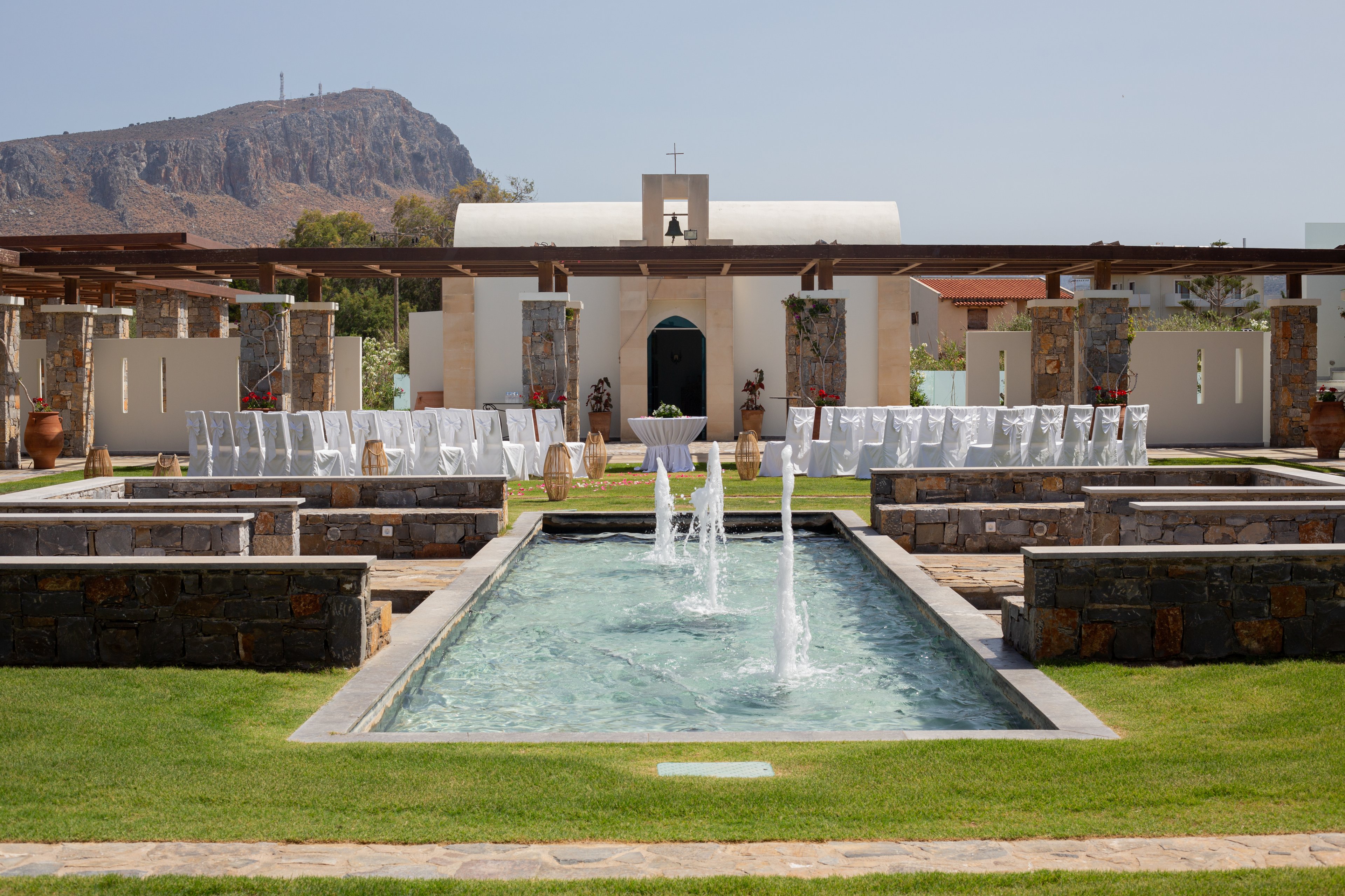 An elegant outdoor wedding ceremony is arranged in front of a small white chapel with a cross on top. Rows of white-covered chairs are lined up on either side of a central aisle facing the chapel entrance. A long rectangular fountain with multiple water jets runs through the middle of the courtyard, framed by stone walls and green lawn. Wooden pergolas, clay pots, and stone columns add rustic architectural details to the scene. A rocky mountain landscape rises in the background under a clear blue sky.