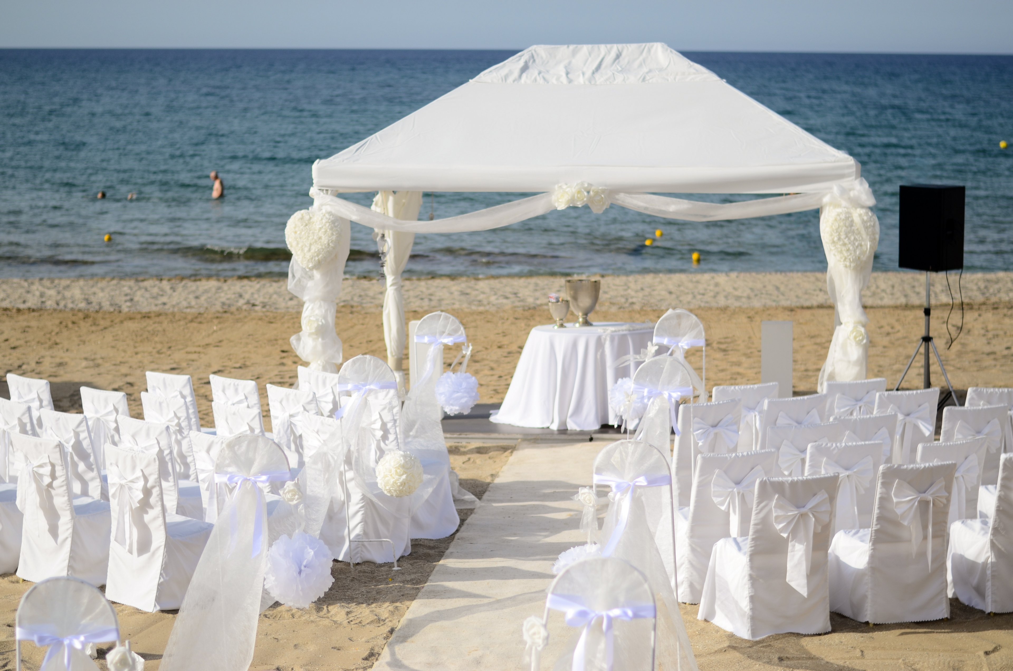 An elegant wedding ceremony setup is arranged on a sandy beach facing the sea. A white canopy with flowing fabric and floral decorations stands at the front, sheltering a small table with ceremonial items. Rows of white chairs with bows line both sides of an aisle runner leading to the altar. The calm ocean and distant swimmers create a serene backdrop under clear skies.