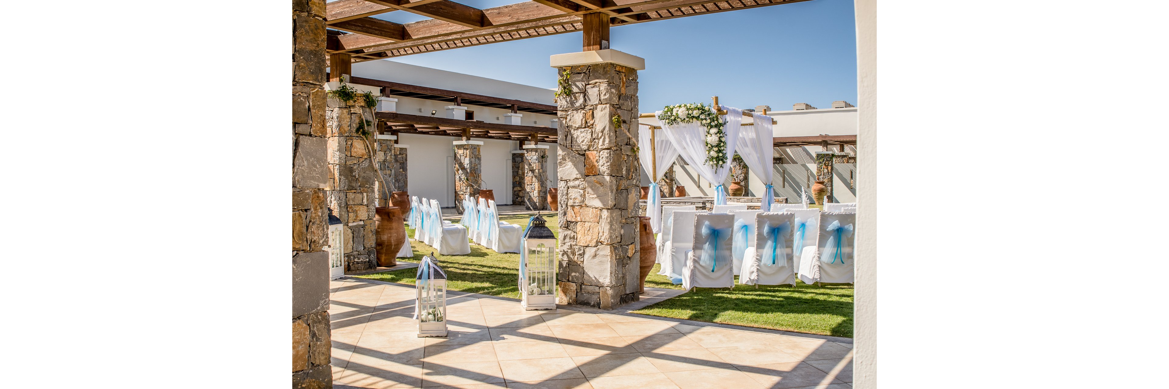 An elegant outdoor wedding ceremony area is arranged beneath a wooden pergola with stone pillars. White chairs decorated with blue ribbons are lined up facing a floral arch draped in white fabric. Large glass lanterns and decorative vases accent the aisle and surrounding space. Bright sunlight and clear blue sky create a warm, festive atmosphere in the courtyard setting.