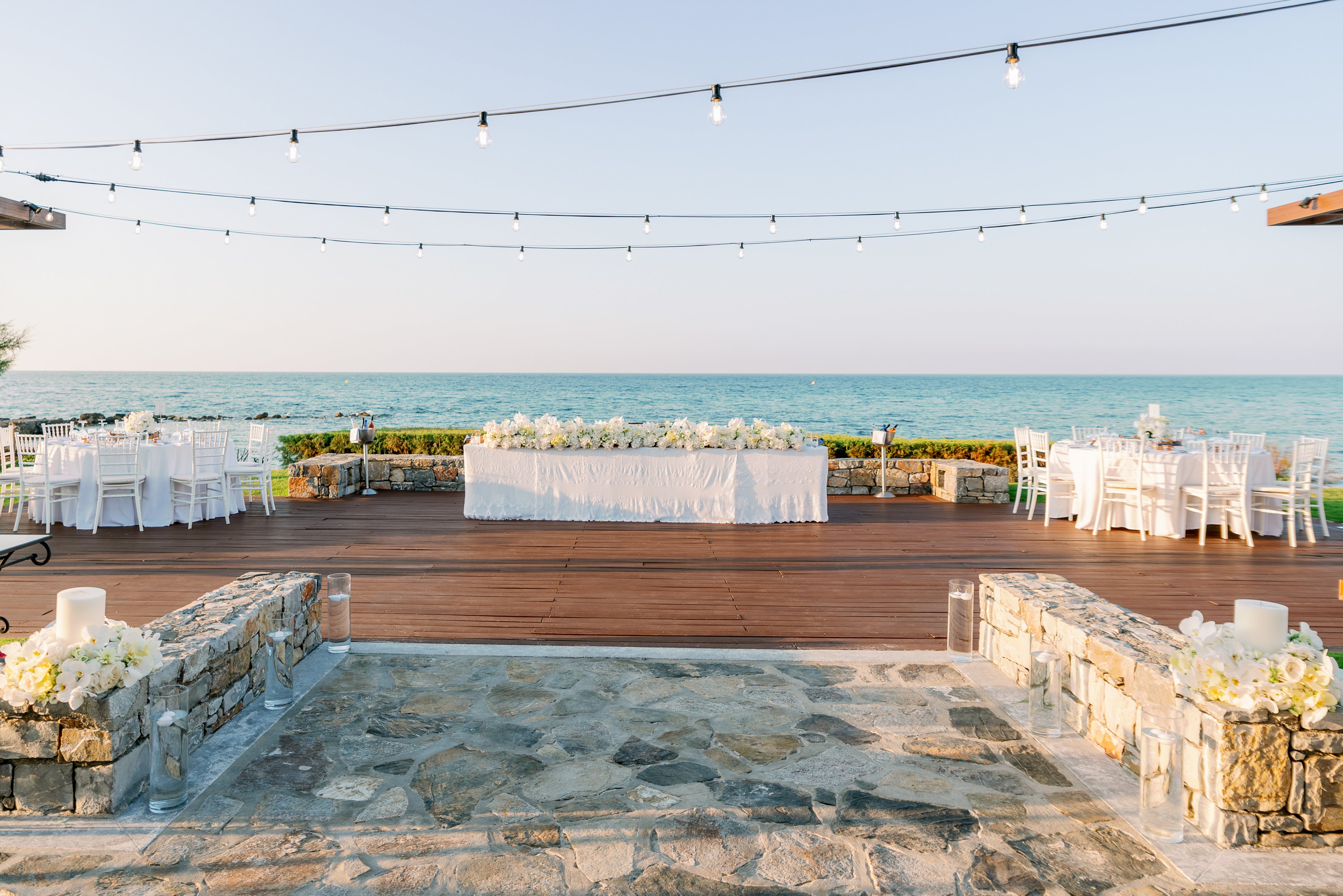 An elegant wedding reception is arranged on a wooden terrace overlooking a calm sea. A long banquet table decorated with white flowers is centered between smaller round guest tables with white chairs. String lights are suspended overhead, adding a festive touch to the open-air setting. Stone steps and low stone walls with floral arrangements frame the entrance to the reception area, creating a romantic coastal atmosphere.