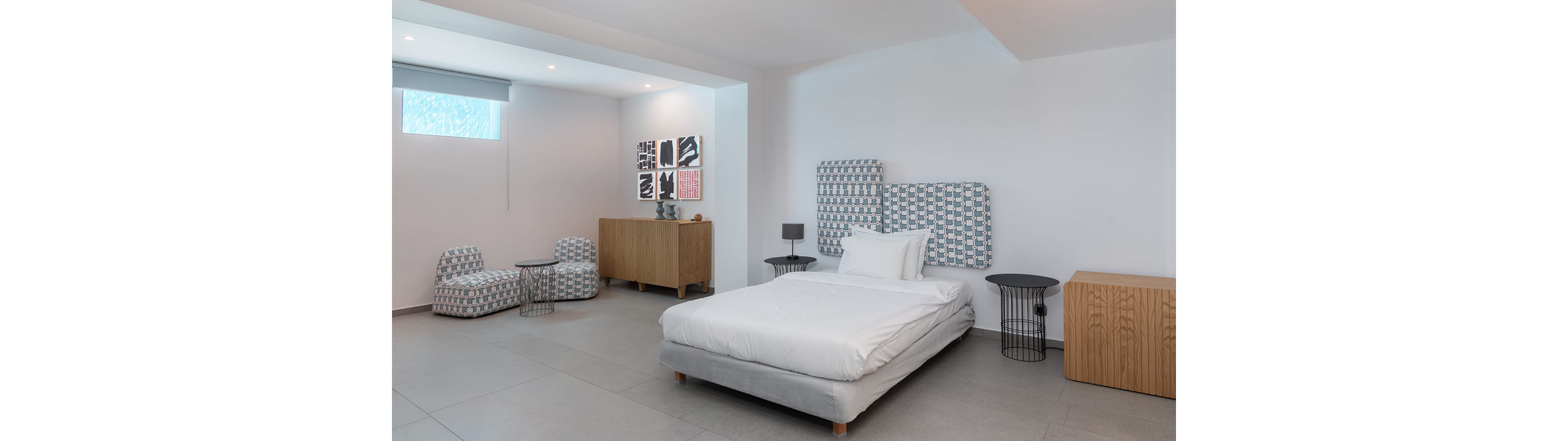 Contemporary bedroom interior featuring a low white bed with patterned headboard and cushions. The room includes matching upholstered lounge chairs, small side tables, and wooden storage cabinets. White walls, large floor tiles, and a high window create a bright, minimalist atmosphere. Abstract black and white artwork adds visual interest to the clean, modern design.