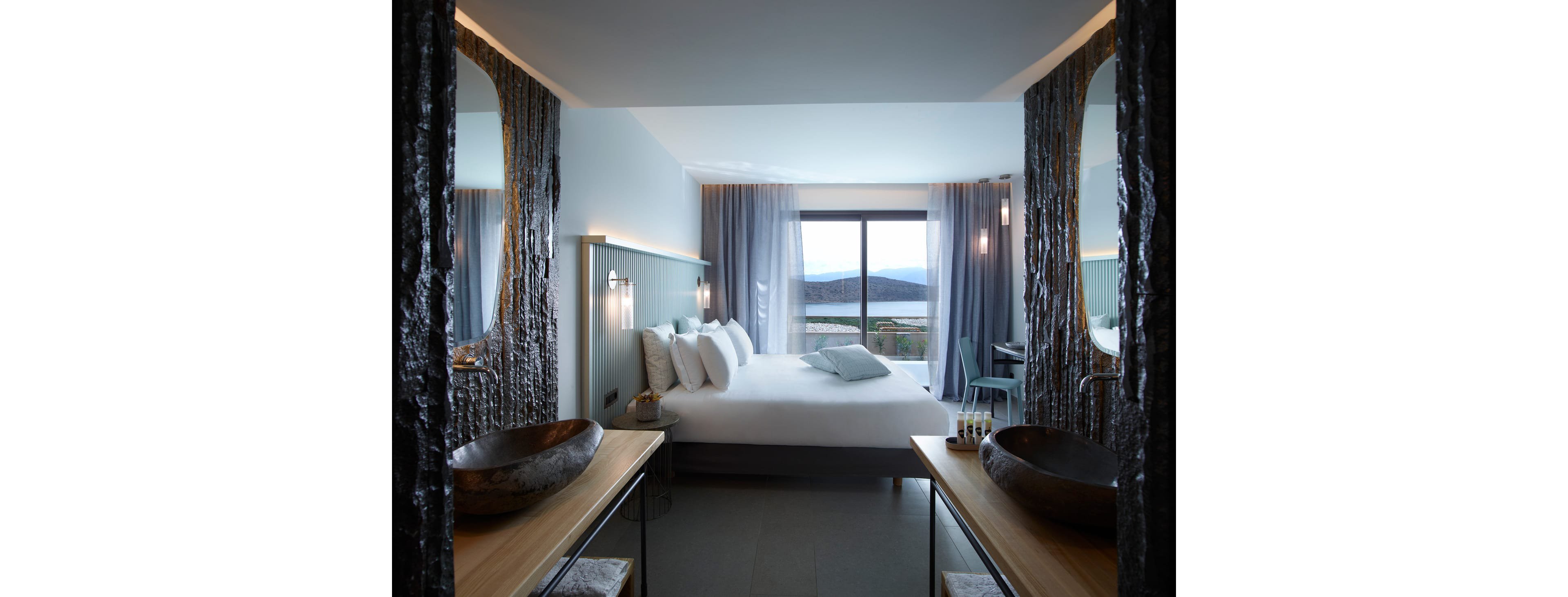 Contemporary hotel bedroom features a large bed with multiple white pillows and soft gray bedding. The room includes two rustic wooden vanities with vessel sinks in the foreground, creating a symmetrical composition. Floor-to-ceiling curtains frame a wide window that overlooks distant mountains and a body of water. Cool tones, textured walls, and subtle lighting give the space a calm, luxurious atmosphere.