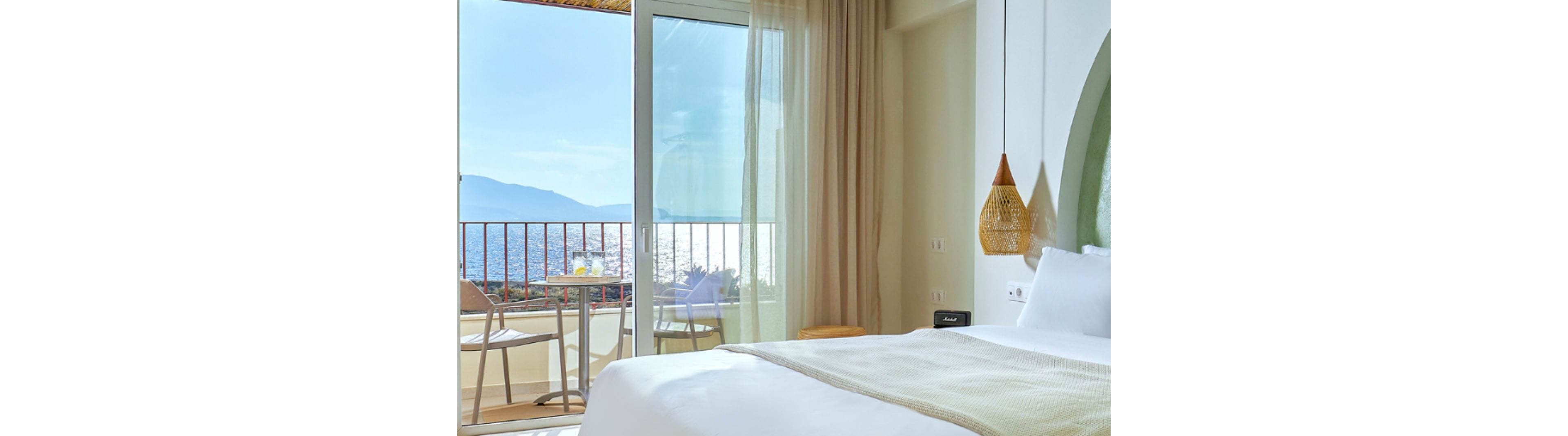 A sunlit hotel bedroom features a neatly made bed with light-colored linens and a woven hanging lamp. Floor-to-ceiling glass doors open onto a balcony overlooking the sea and distant mountains. Outside, a small table and chairs are set up for enjoying the coastal view. Soft neutral tones and natural textures create a calm, relaxing atmosphere.
