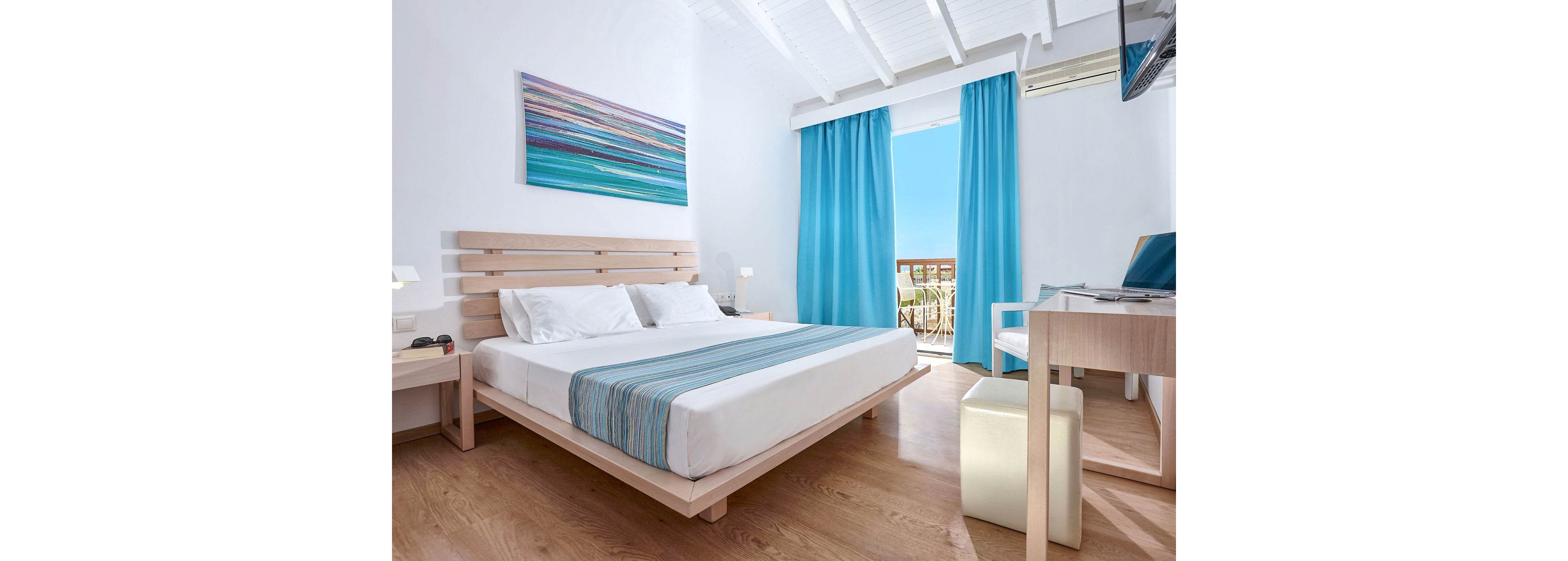 Modern hotel bedroom with a double bed features light wood furniture and white walls. A striped blue bed runner and matching curtains frame a balcony with a sunny sky and outdoor seating. The room includes a desk with chair, bedside tables, and a wall-mounted television. Overall, the space feels airy, minimalist, and inspired by coastal colors.