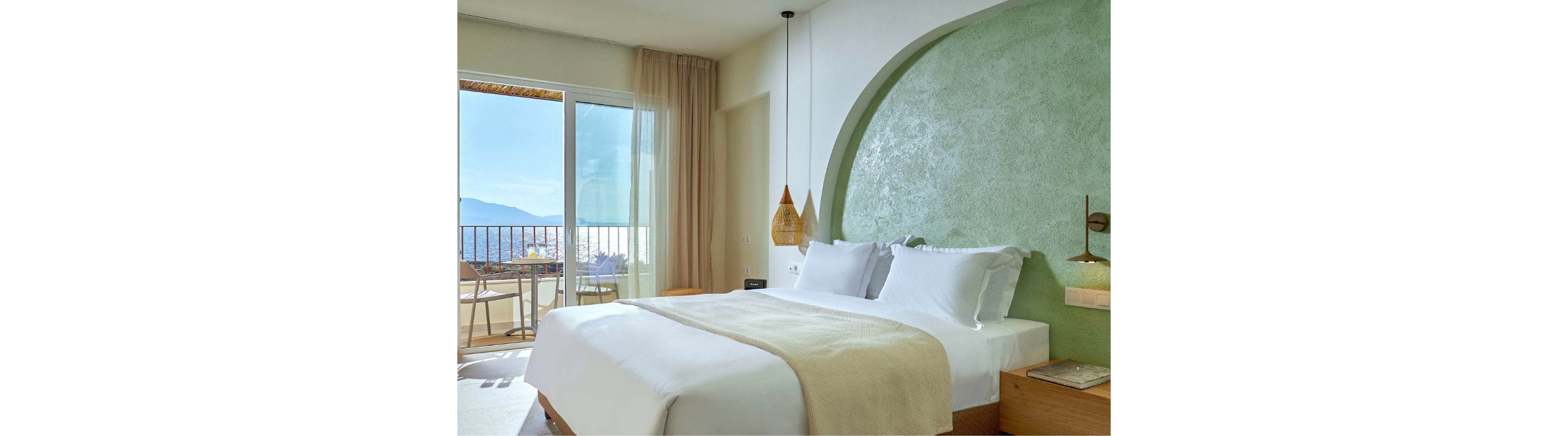 Bright contemporary hotel bedroom with a large neatly made bed and white linens. A textured green accent wall curves behind the headboard, complemented by wooden bedside tables and a woven pendant light. Floor-to-ceiling curtains frame sliding glass doors that open to a balcony with chairs overlooking the sea and distant mountains. Soft natural light creates a calm, relaxing atmosphere ideal for a beachside retreat.