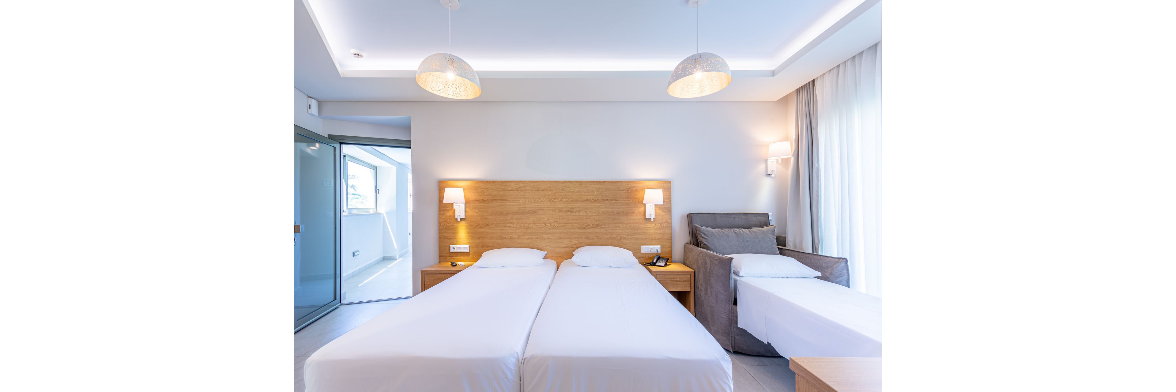 A bright, modern hotel room features two neatly made twin beds with white linens and a wooden headboard. Soft wall-mounted lamps and ceiling lights create a warm, inviting atmosphere. A gray armchair and an additional single bed are placed to the side near a large window with sheer curtains. A glass door opens to an adjacent balcony or sunroom, enhancing the airy, minimalist design.