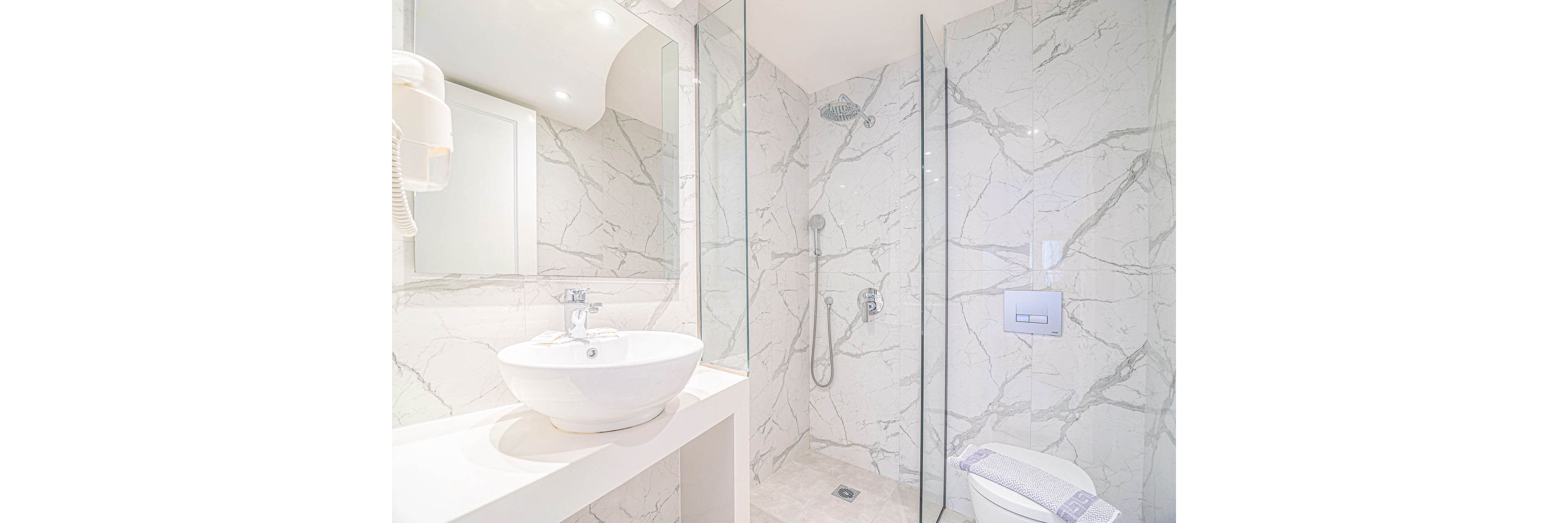 A bright modern bathroom features a walk-in shower with glass walls and a rainfall showerhead. The walls and floor are covered in white marble tiles with gray veining, creating a clean and luxurious look. A round vessel sink sits on a sleek countertop beside a wall-mounted mirror and hair dryer. A compact toilet with a folded towel on top completes the minimalist, spa-like space.