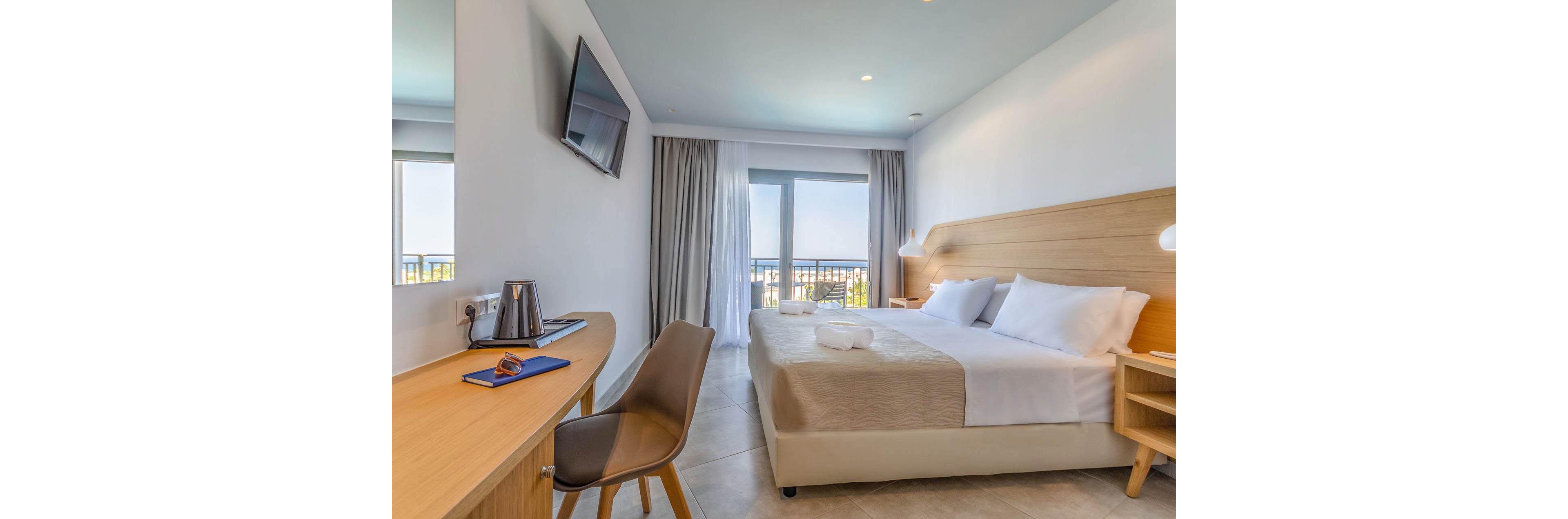A contemporary hotel room features a neatly made double bed with white linens and a light wood headboard. A wall-mounted flat-screen TV faces the bed, and a minimalist desk with a chair, kettle, and notebook sits to the side. Floor-to-ceiling curtains frame a glass door leading to a balcony with a scenic outdoor view. Soft neutral tones, clean lines, and ample natural light create a calm, inviting atmosphere.