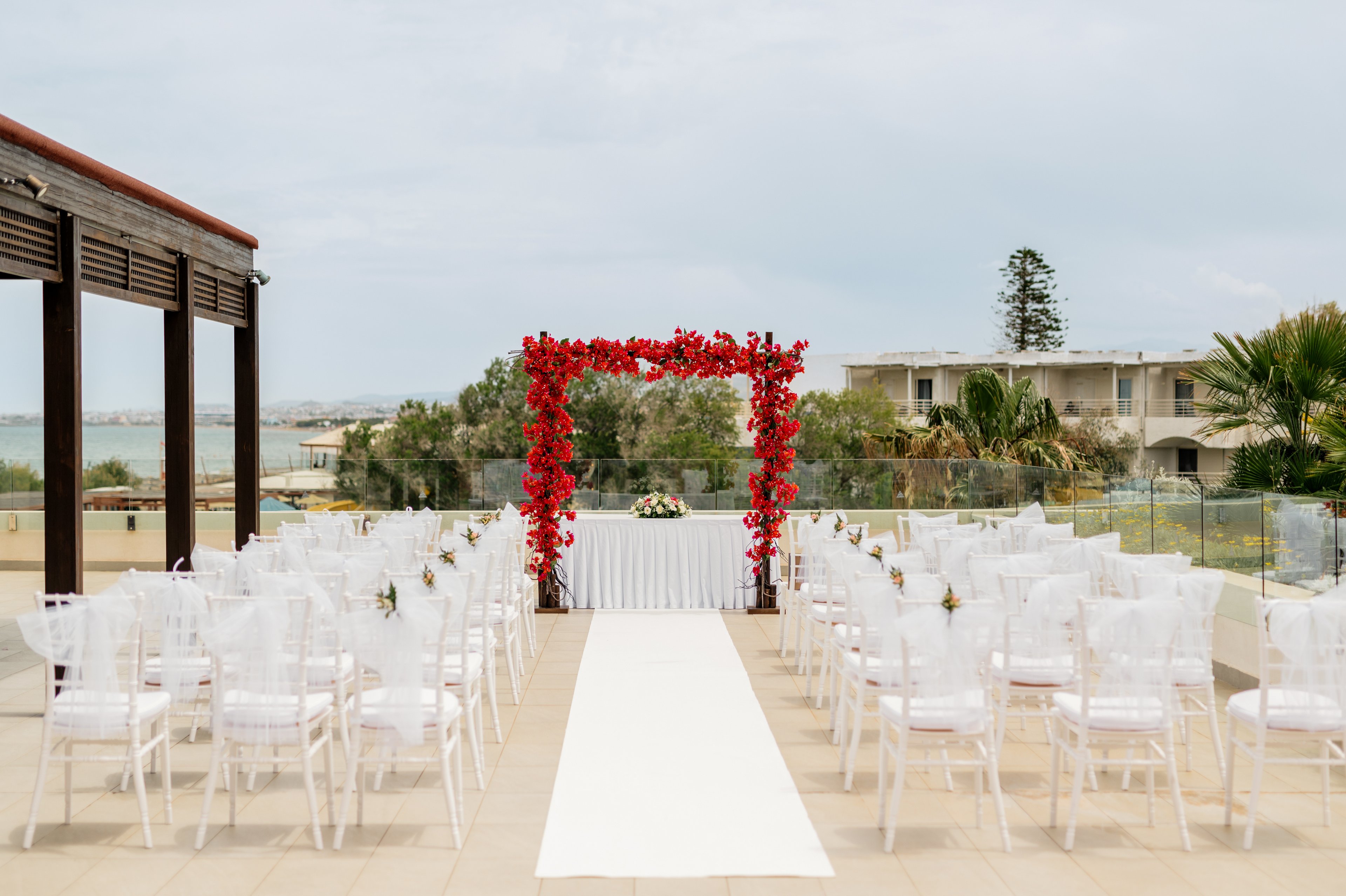 An outdoor wedding ceremony area is arranged with rows of white chairs facing a floral arch. The arch is decorated with vibrant red flowers and frames a small table at the front. A white aisle runner leads from the foreground to the altar, creating a clean, elegant path. The venue overlooks a coastal landscape with buildings, trees, and calm water in the background under a cloudy sky.
