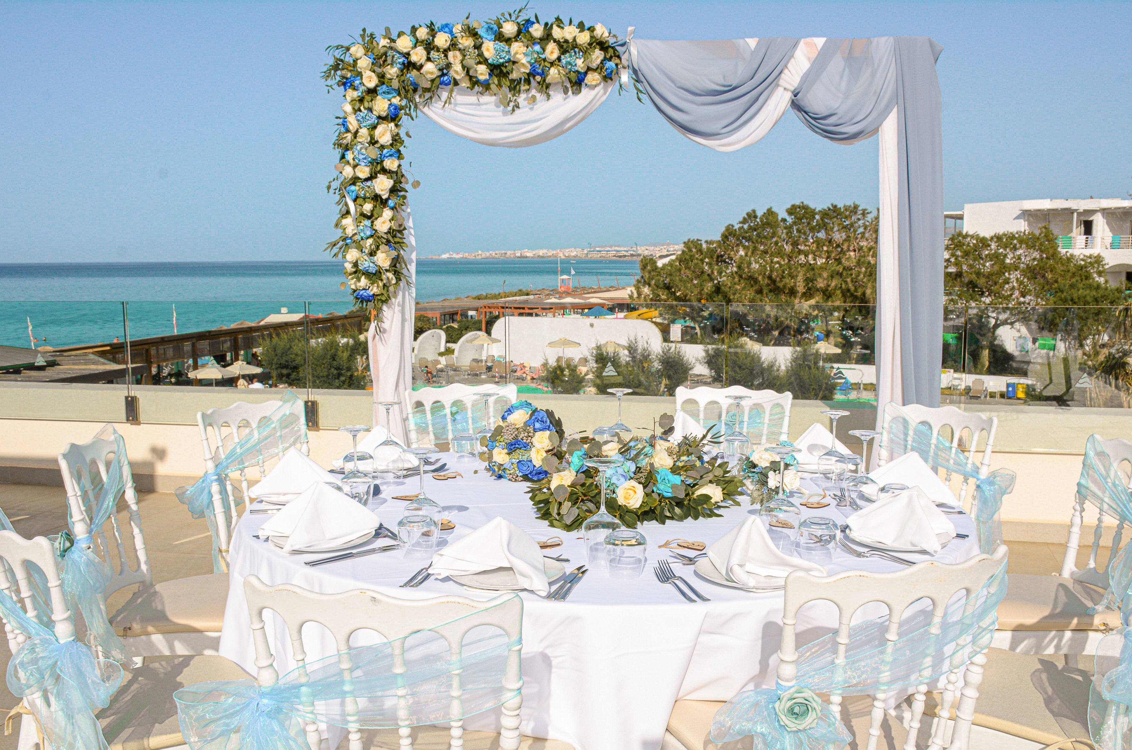 A round banquet table is beautifully set for a wedding reception on a terrace overlooking the sea. White chairs with light blue tulle bows surround the table, which is arranged with folded napkins, glassware, and floral centerpieces. An arch draped in white fabric and decorated with blue and white flowers frames the view of the coastline. The bright daylight and clear blue water create a fresh, romantic atmosphere.