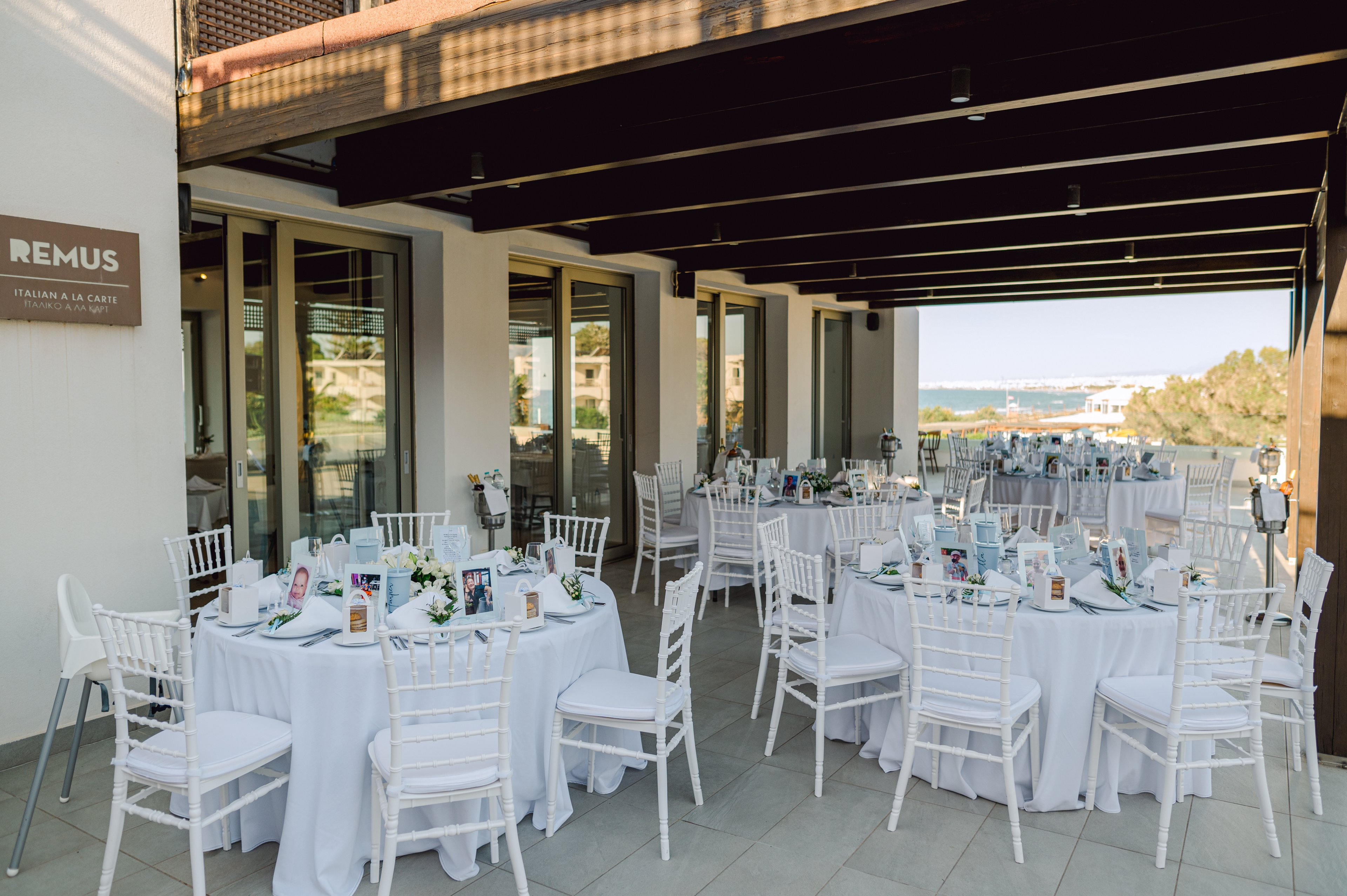 Round tables with white linens and white chairs are arranged on a covered outdoor terrace, prepared for a formal event or reception. Each table is decorated with small centerpieces and printed table numbers or photos. Large glass doors reflect the bright daylight, while the open side of the terrace reveals a distant view of the sea and coastline. A wall sign reading "REMUS" and additional text indicates the venue name near the entrance.