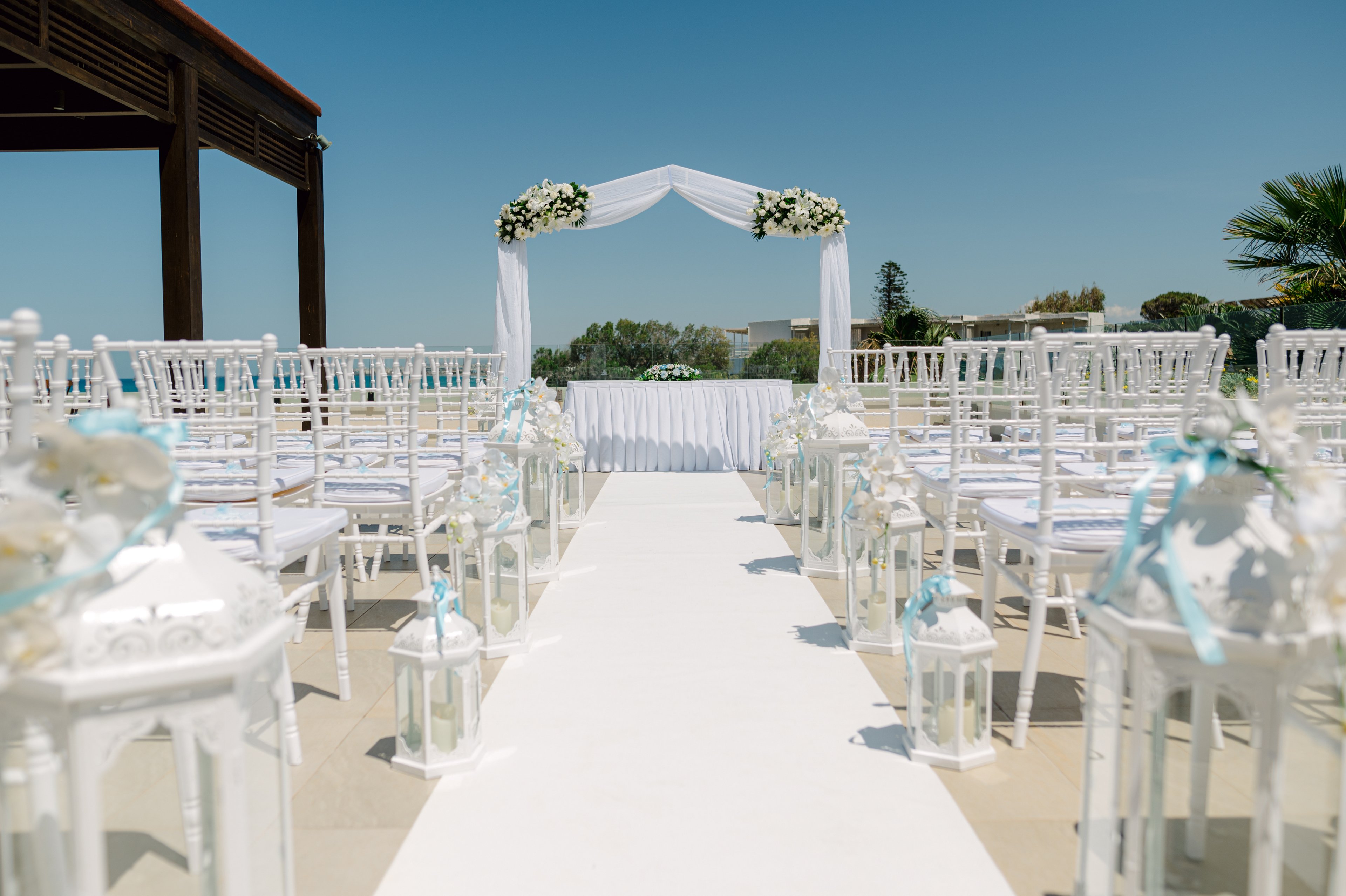 An outdoor wedding ceremony setup features a white aisle leading to a floral arch. Rows of white chairs line both sides of the aisle, decorated with light blue ribbons and white flowers. White lanterns are placed along the walkway, enhancing the elegant and romantic atmosphere. A clear blue sky and distant greenery create a bright, summery backdrop.