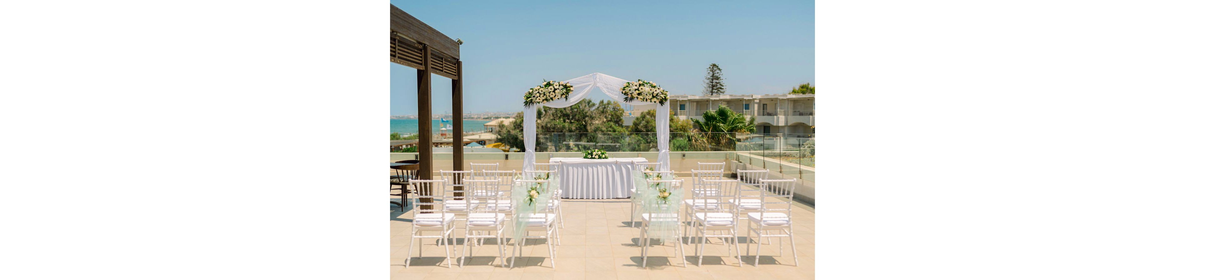 An elegant outdoor wedding ceremony setup is arranged on a terrace overlooking the sea. White chairs are lined in rows facing a floral-decorated arch and a table draped in white cloth. Soft green and white floral arrangements adorn the chairs and the arch, creating a romantic atmosphere. Modern buildings, glass railings, and a clear blue sky frame the scenic coastal backdrop.