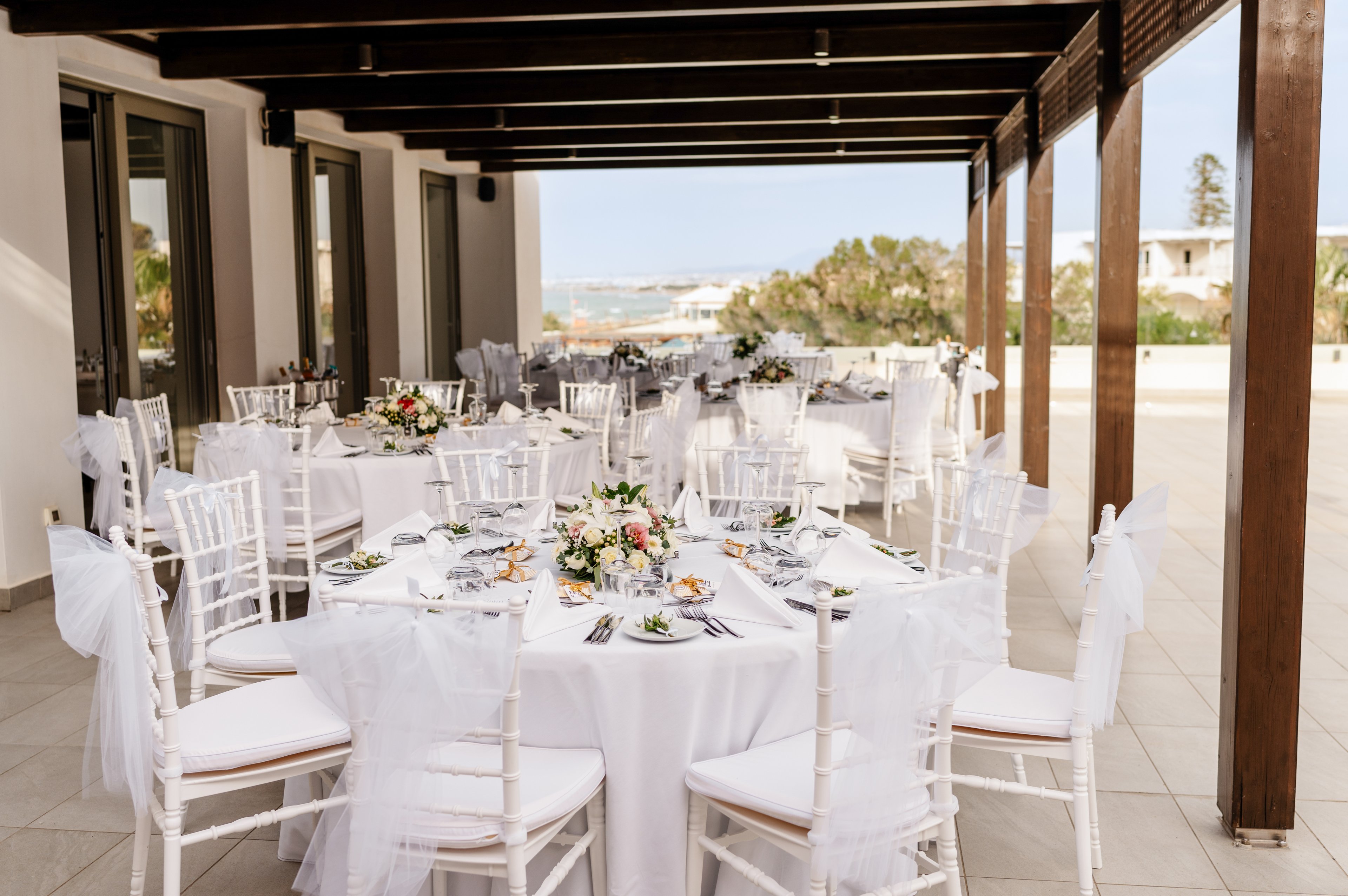Round tables with white linens and floral centerpieces are arranged on a covered terrace for an outdoor wedding reception. White chiavari chairs with sheer fabric bows surround each table, creating a refined and romantic atmosphere. The venue overlooks a coastal landscape with sea and distant buildings visible in the background. Soft natural light enhances the clean, airy feel of the celebration setting.