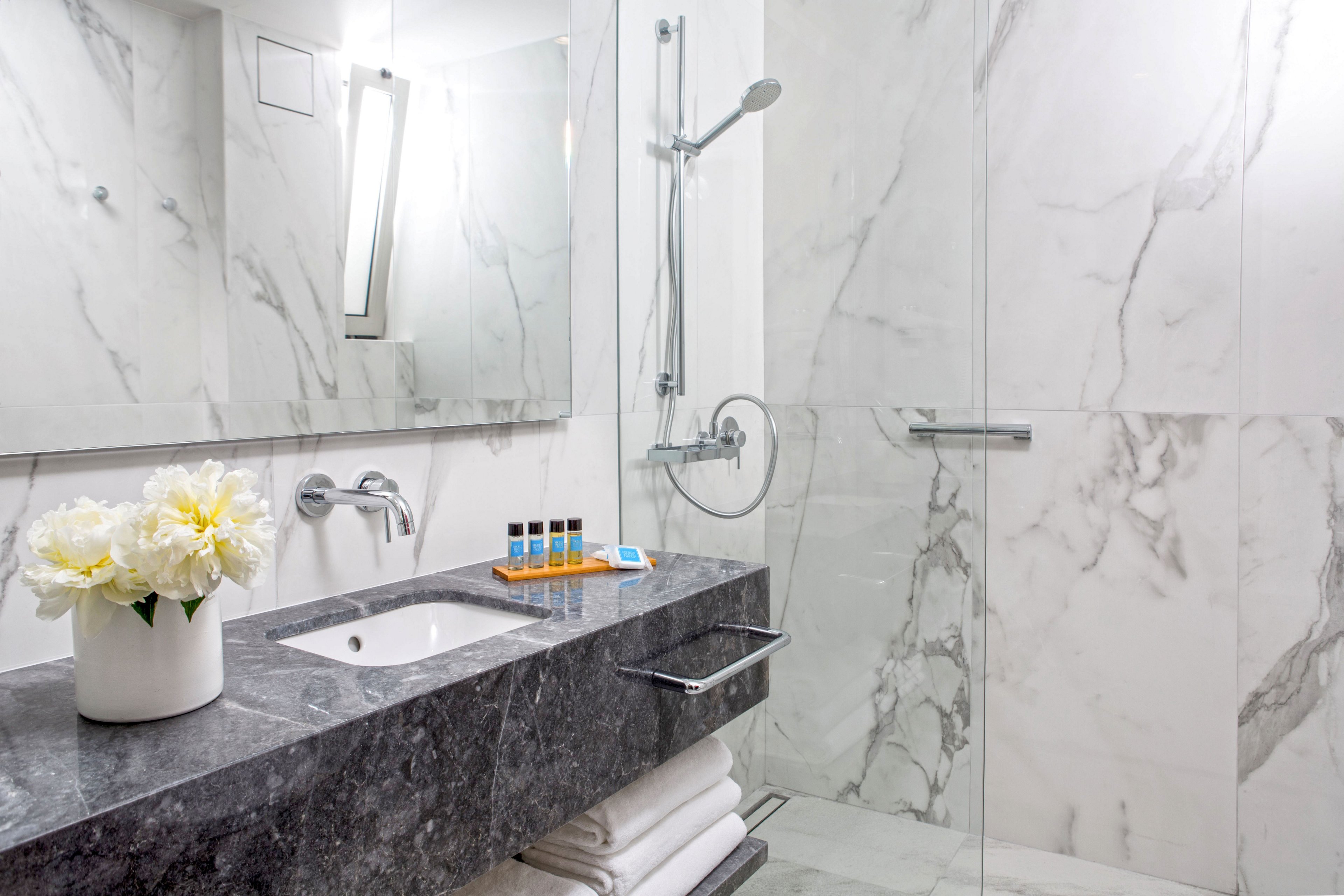 A contemporary bathroom features a sleek gray stone vanity against white marble walls. A glass-enclosed walk-in shower with a handheld showerhead and linear drain is visible to the right. On the countertop, a tray holds small toiletry bottles beside a sink with a minimalist faucet, while white towels are neatly stacked underneath. A vase of white flowers and a vertical wall light add a touch of elegance and warmth to the clean, modern space.
