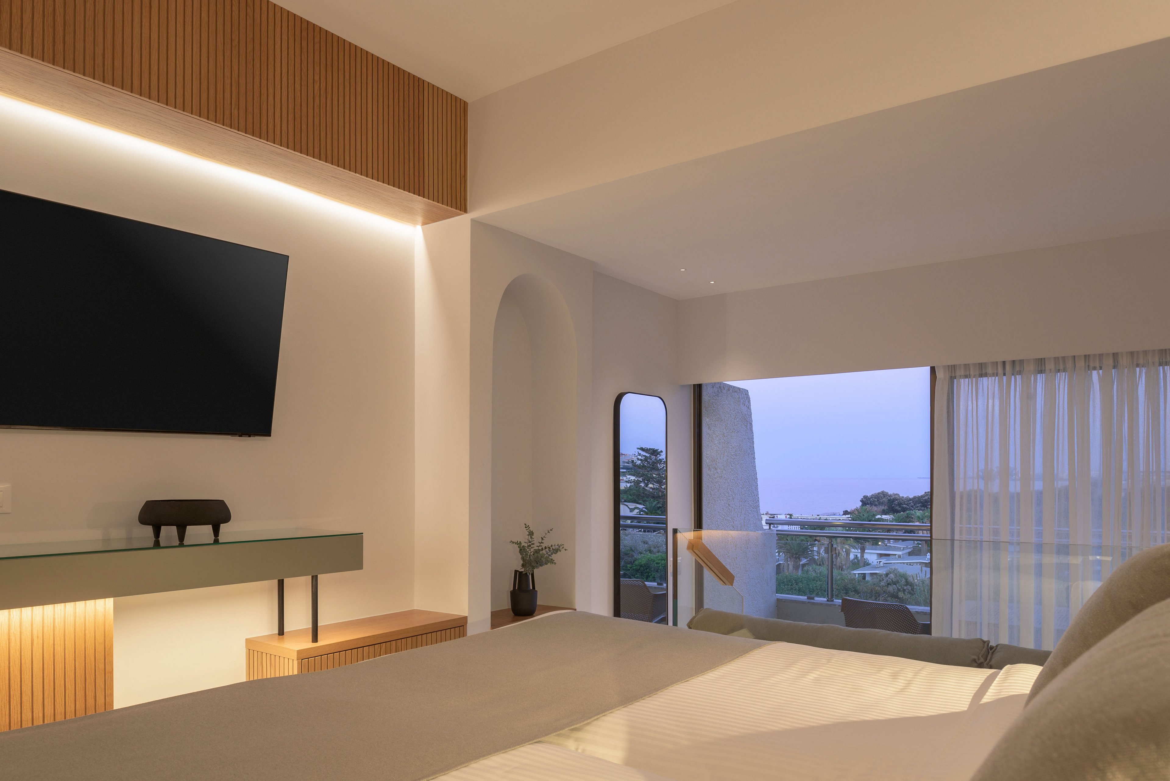 A contemporary hotel bedroom features a large bed facing a wall-mounted flat-screen TV and minimalist console. Warm indirect lighting and wood accents create a cozy, modern atmosphere. A glass door opens to a balcony with outdoor seating and views of trees and distant water at dusk. Sheer curtains and soft neutral tones enhance the calm, relaxing setting.
