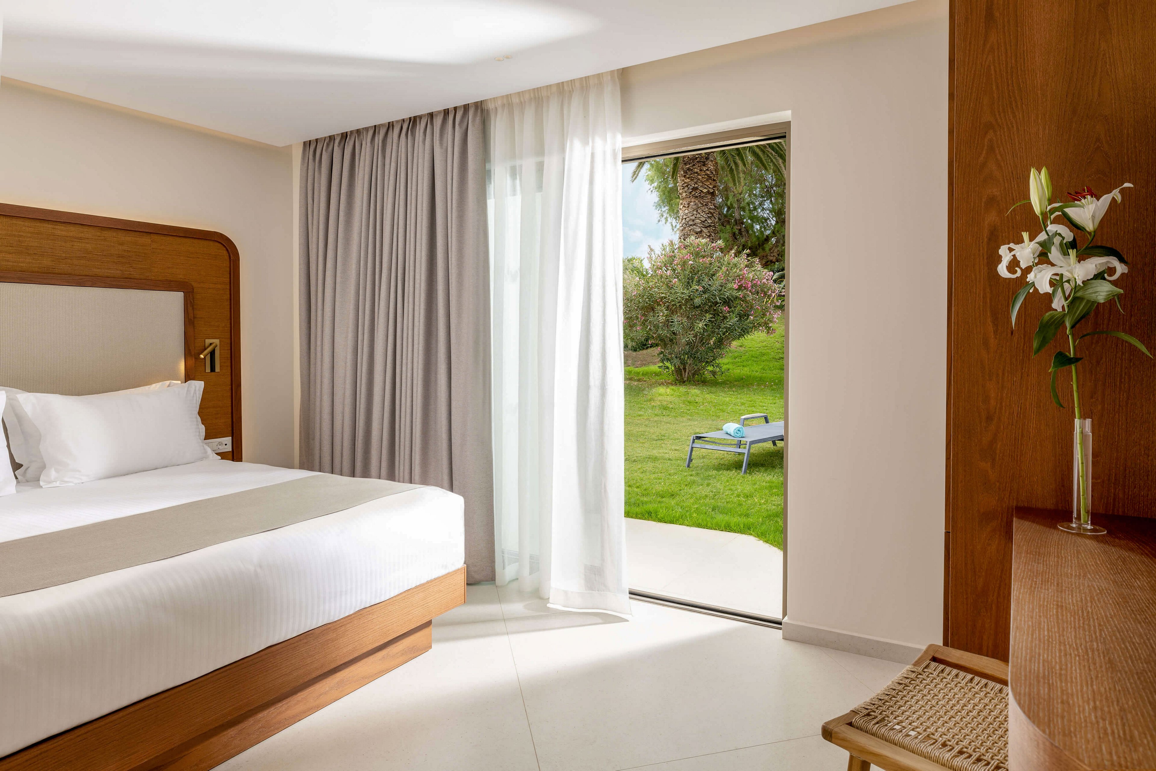 A modern hotel room features a neatly made double bed with crisp white linens and a wooden headboard. Floor-to-ceiling curtains frame a sliding glass door that opens directly onto a lush green garden. Outside, a lounge chair sits on the grass near palm trees and flowering shrubs. Warm neutral tones, natural light, and minimalist decor create a calm, inviting atmosphere.