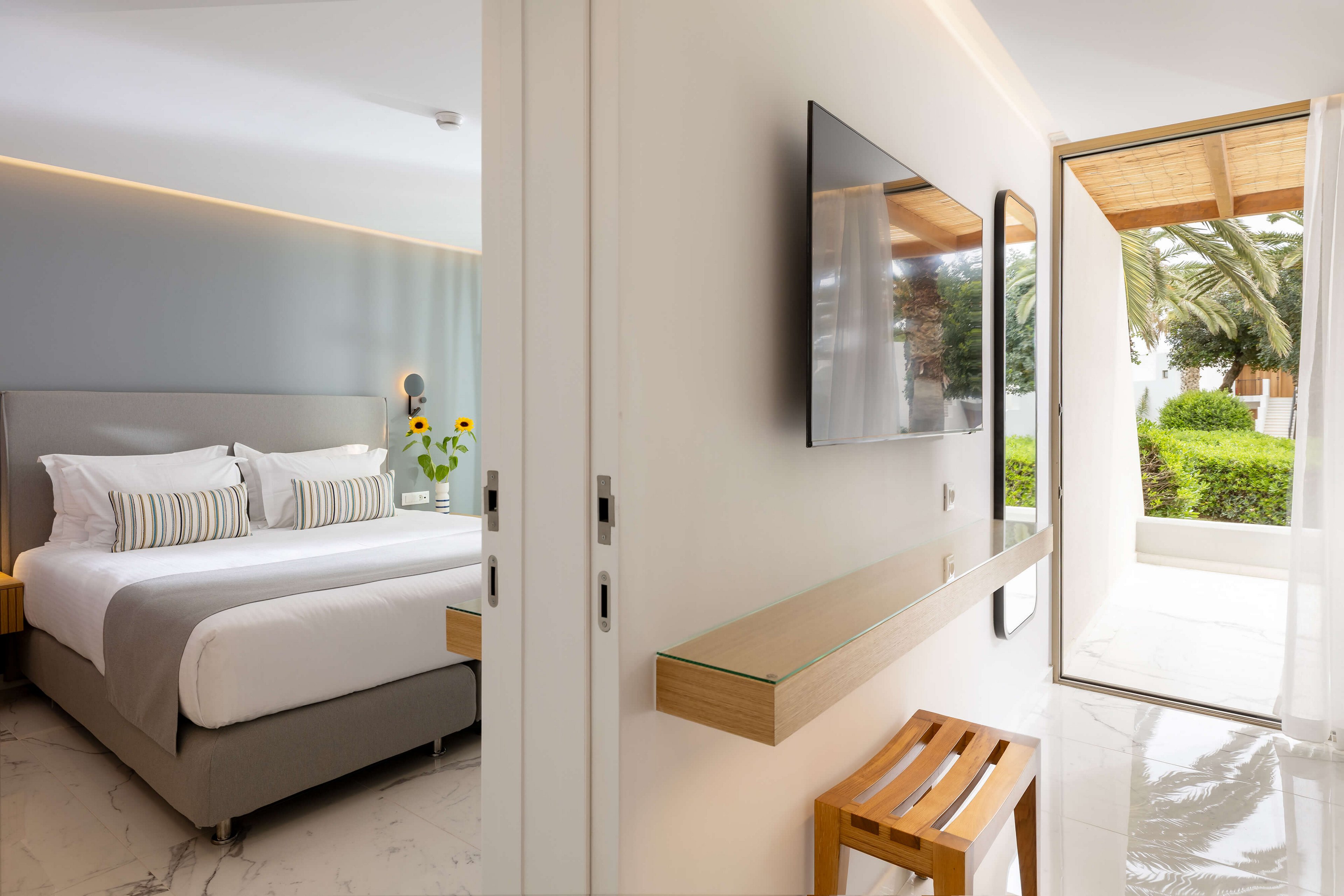 A contemporary hotel bedroom features a large neatly made bed with striped pillows and a soft gray headboard. The room opens onto a bright terrace with greenery and palm trees visible outside. A wall-mounted flat-screen television and a minimalist glass shelf with a wooden stool enhance the clean, modern design. Natural light floods the space, highlighting the white marble-style floor and calm neutral color palette.