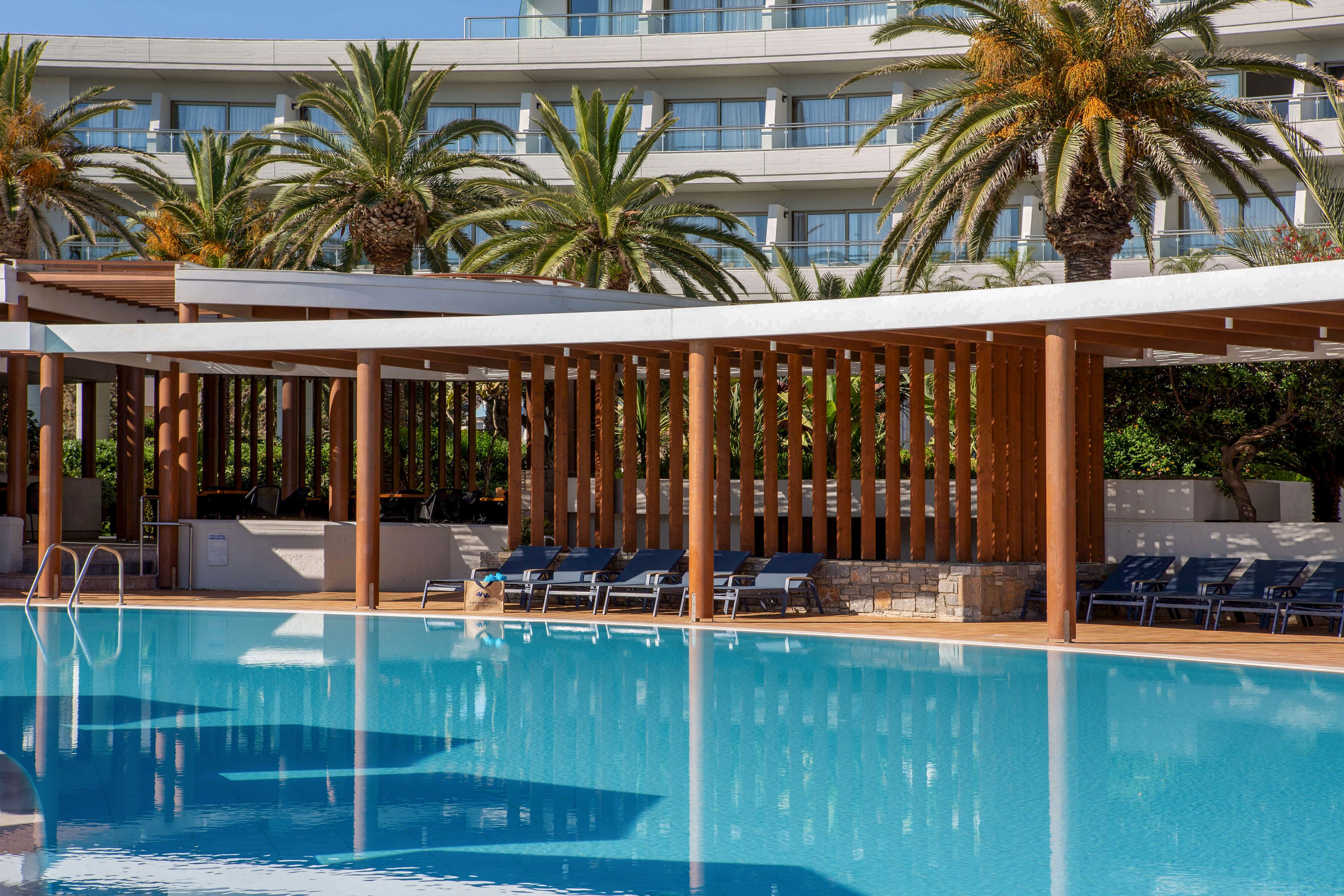 A large, clear blue swimming pool sits in front of a modern resort structure with wooden slats and white canopy roofs. Several lounge chairs line the shaded poolside area, suggesting a relaxing vacation setting. Tall palm trees rise behind the structure, framing the contemporary hotel building in the background. The scene is bright and sunny, emphasizing leisure and outdoor relaxation.