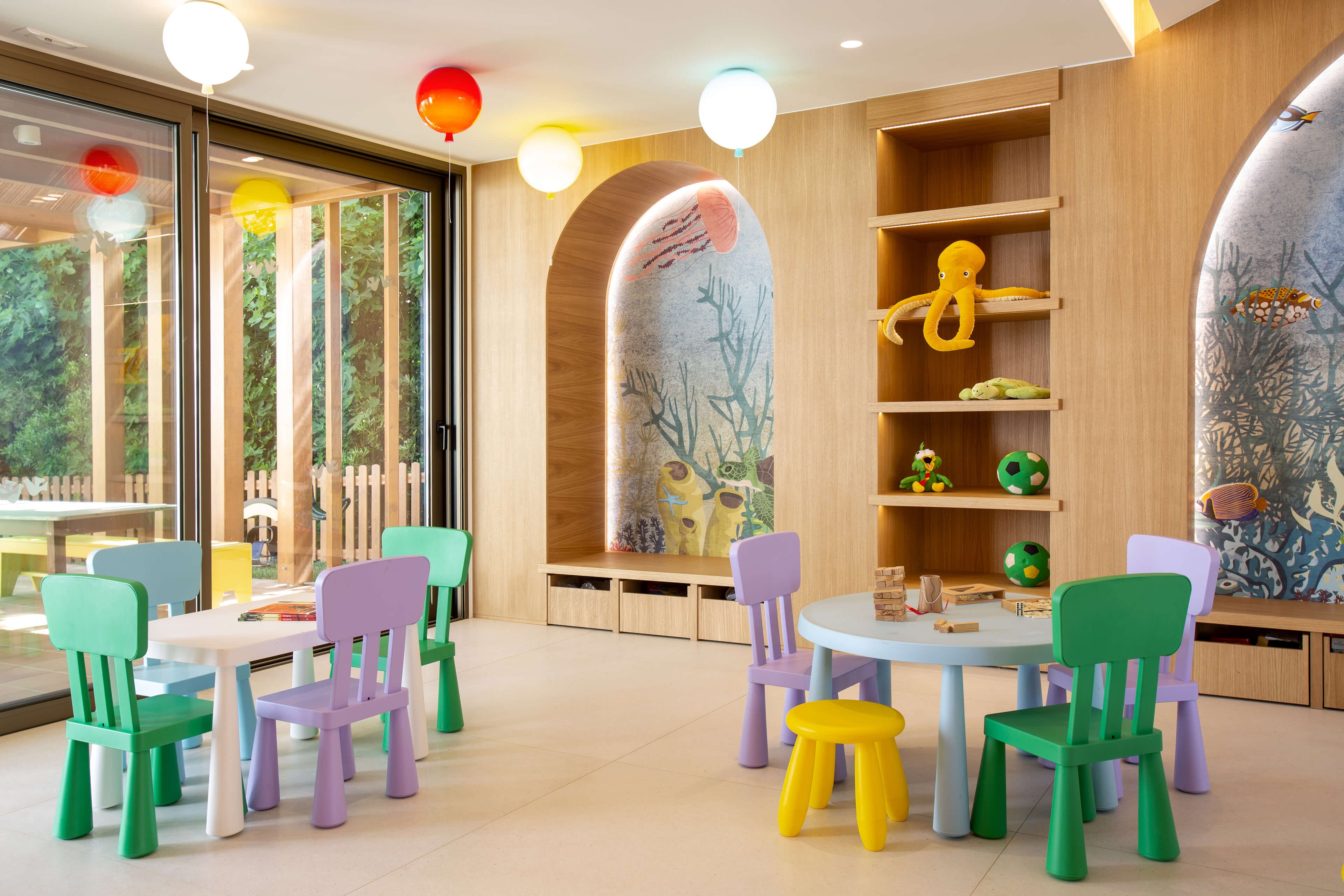 A bright children’s playroom features small round tables surrounded by colorful plastic chairs. Wooden shelves and arched wall niches display ocean-themed decorations, including fish illustrations and a large yellow octopus figure. Large windows open to a green outdoor area, filling the room with natural light. Hanging globe lights and pastel furniture create a cheerful, inviting atmosphere for kids’ activities.