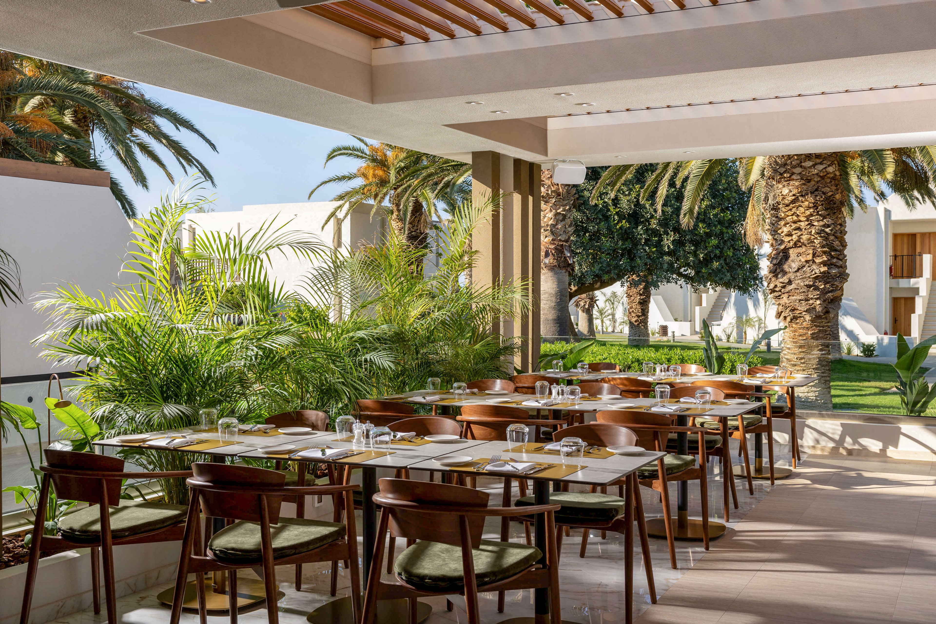 An open-air restaurant terrace features neatly arranged wooden tables and chairs. Each table is set with glassware, plates, and napkins, ready for guests. Lush green plants and tall palm trees surround the seating area, creating a tropical garden atmosphere. Sunlight filters through the slatted roof, casting soft shadows and enhancing the relaxed, resort-like setting.