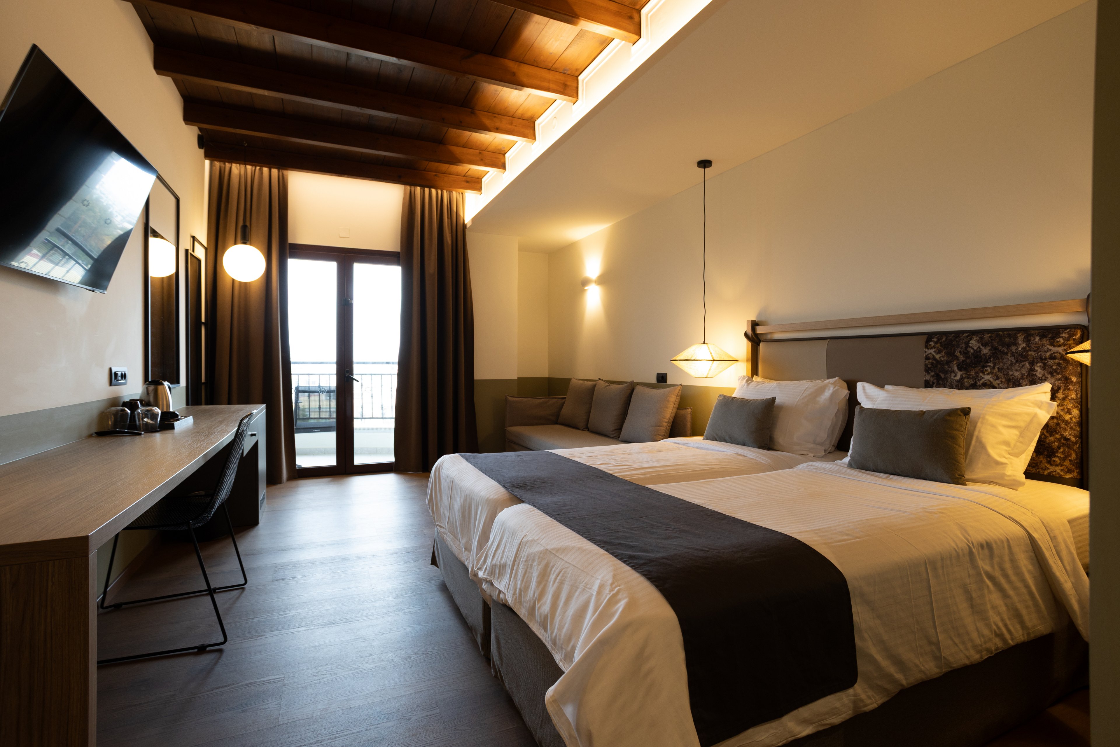 A contemporary hotel room features a large neatly made bed with white linens and dark accent runners. The space includes a wooden desk with a chair, a wall-mounted flat-screen television, and soft ambient lighting from pendant and wall lamps. Floor-to-ceiling curtains frame a glass door that opens to a balcony, allowing natural light into the room. Exposed wooden ceiling beams and neutral tones create a cozy yet modern atmosphere.