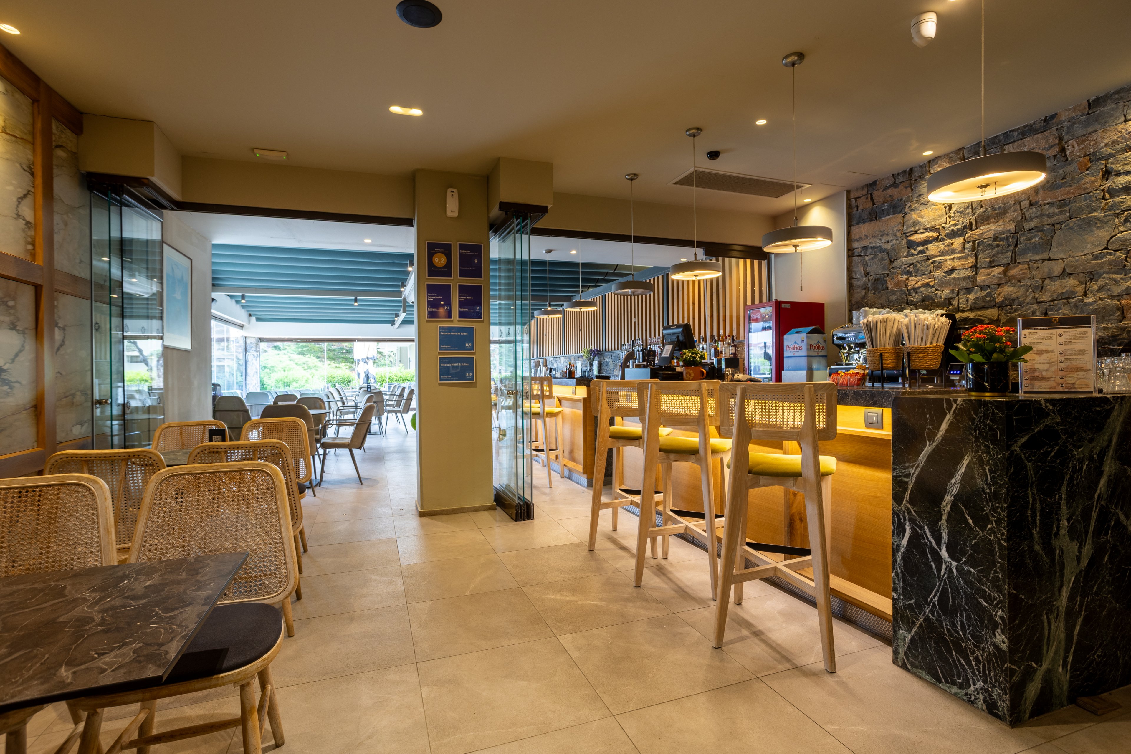 A contemporary café interior features a long bar counter with high wooden stools and a dark marble front. Wicker chairs and small tables fill the seating area, leading toward large open windows that reveal greenery outside. Warm pendant lights and recessed ceiling lights create a welcoming atmosphere, highlighting stone and wood textures on the walls. The space appears calm and inviting, suitable for casual dining or coffee breaks.